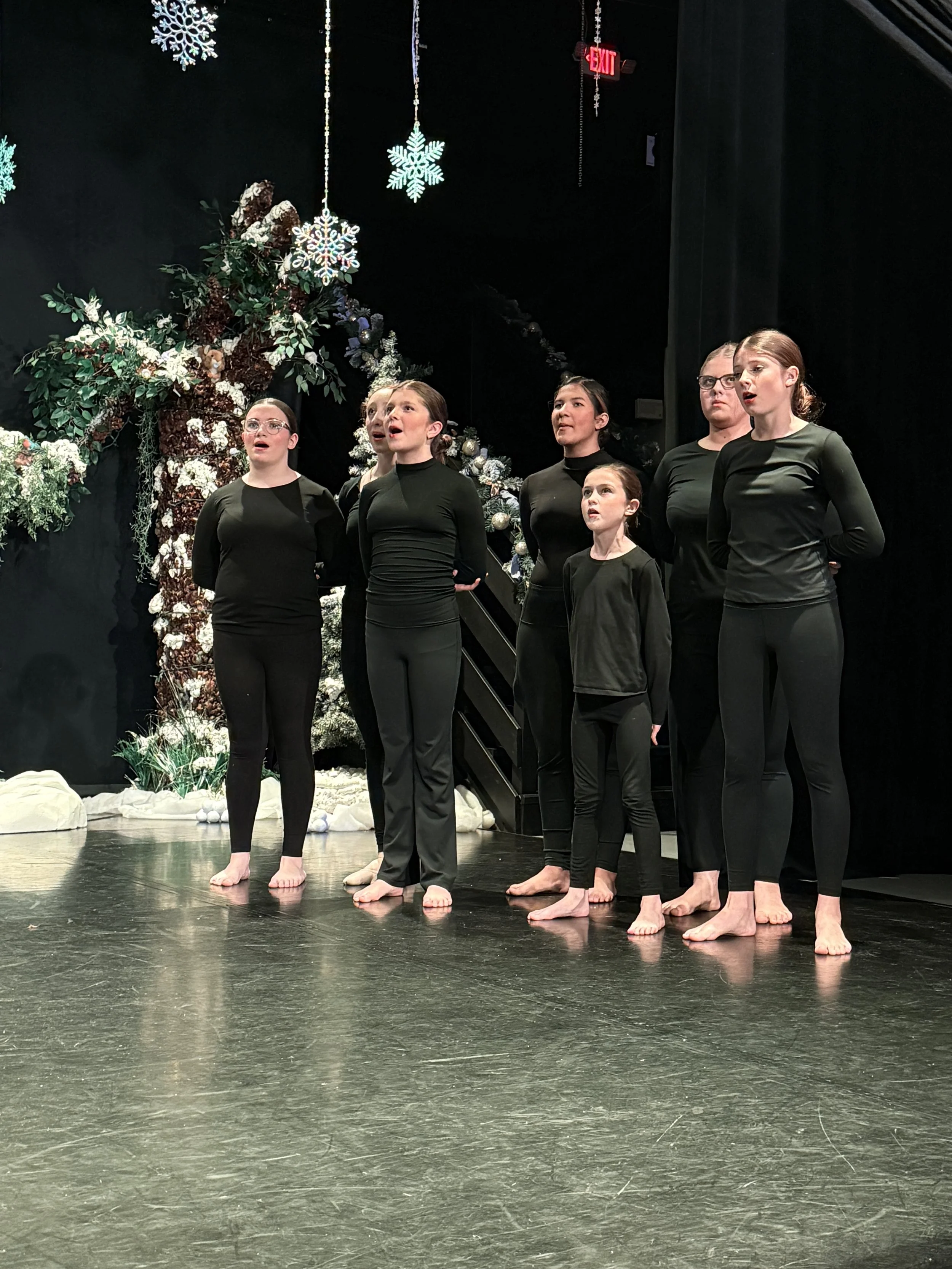 Group of six young girls singing on stage during a holiday performance with winter-themed decorations, snowflakes, and a decorated tree in the background.