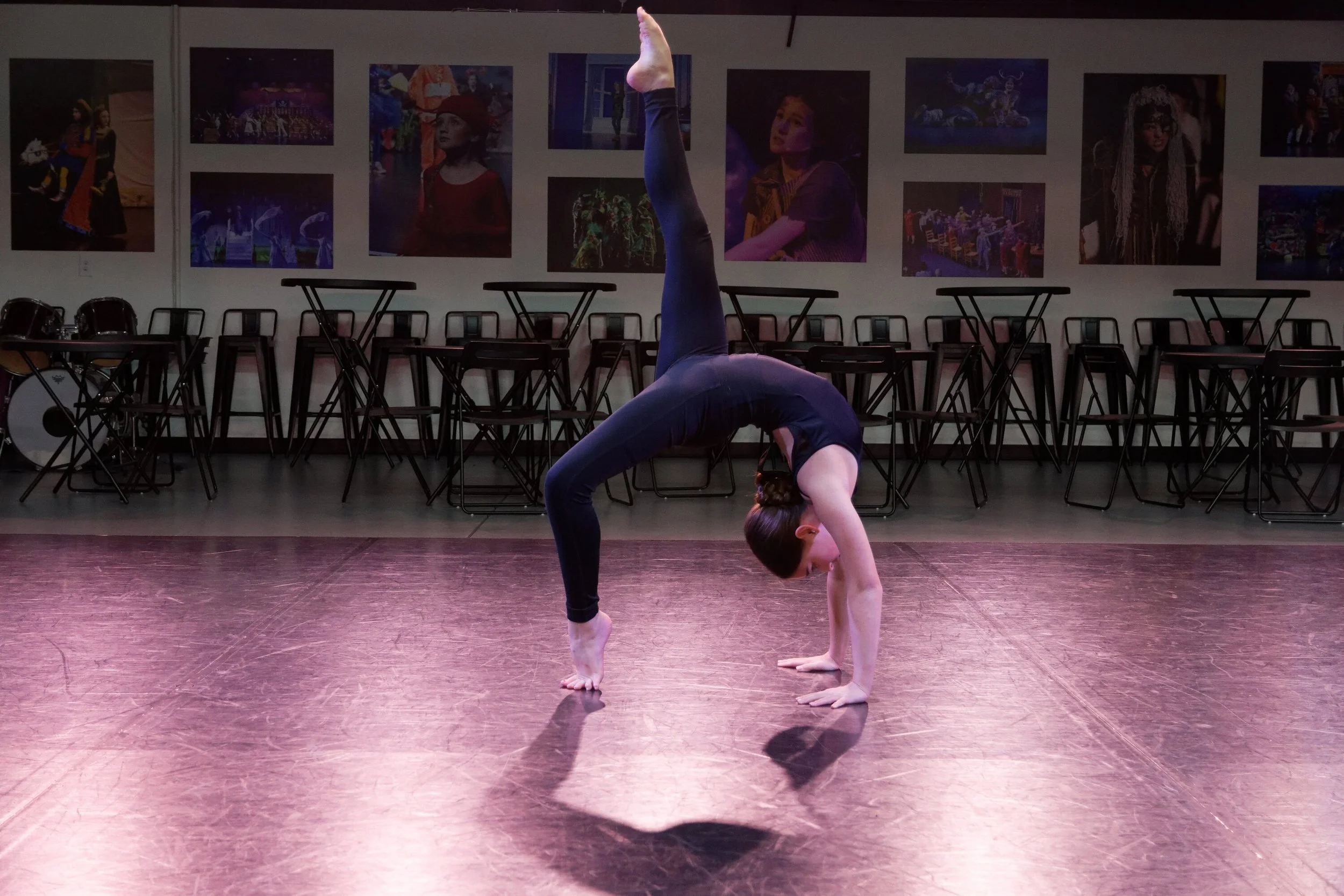 A young girl practicing a handstand in a dance studio with pink reflective floor, black walls, chairs, tables, and colorful theatrical posters on the wall.