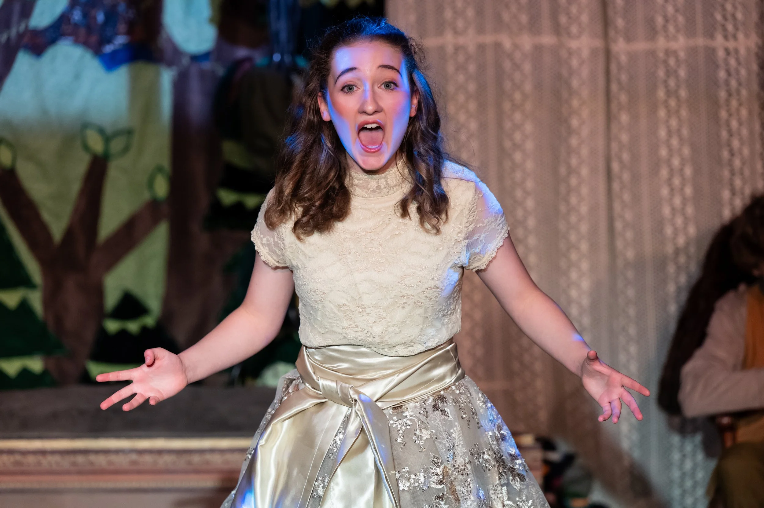 Young girl with shoulder-length curly hair, wearing a cream-colored lace top and a metallic gold skirt, with her mouth open and arms outstretched, appears to be shouting or singing on stage.