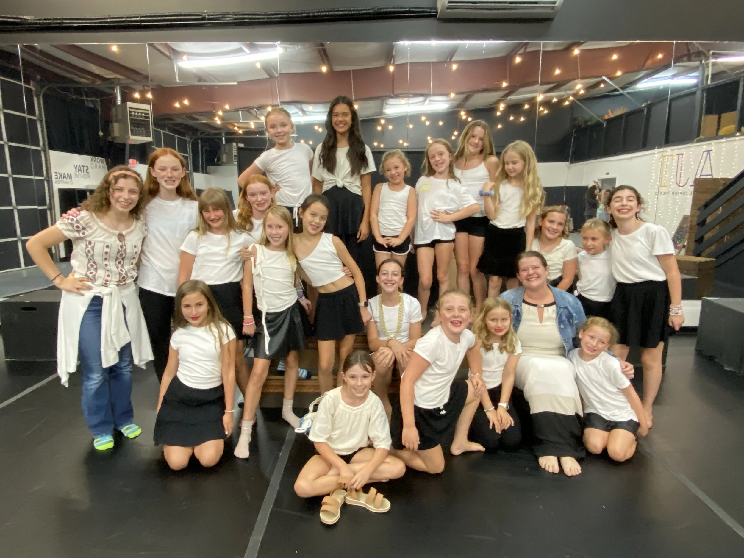 A group of girls and two women, some wearing white t-shirts and black skirts or shorts, posing on stage in a black and white dance studio with string lights overhead.