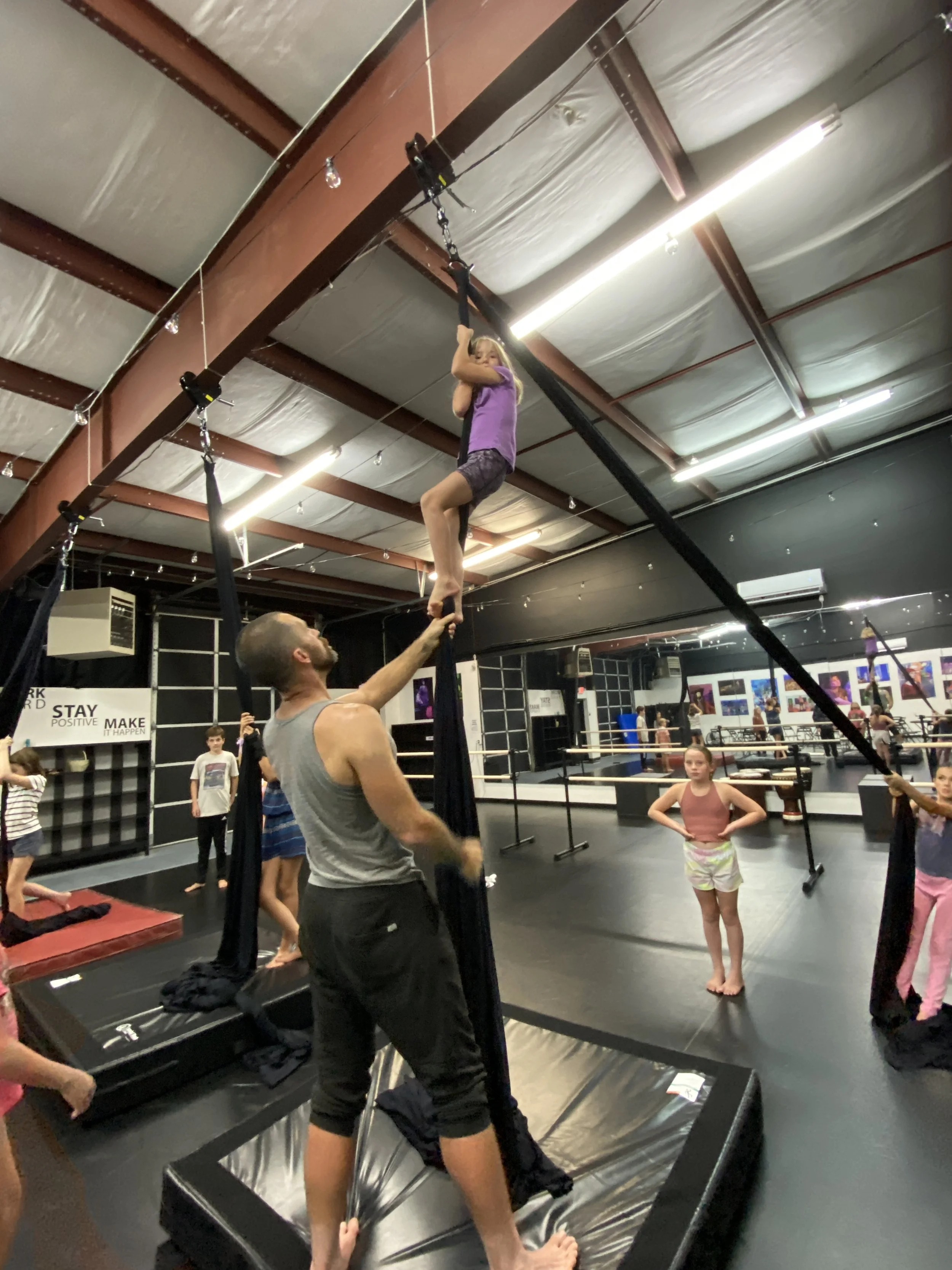 Young girl practicing aerial silks in a dance or gymnastics studio, assisted by a male instructor, with other children observing.