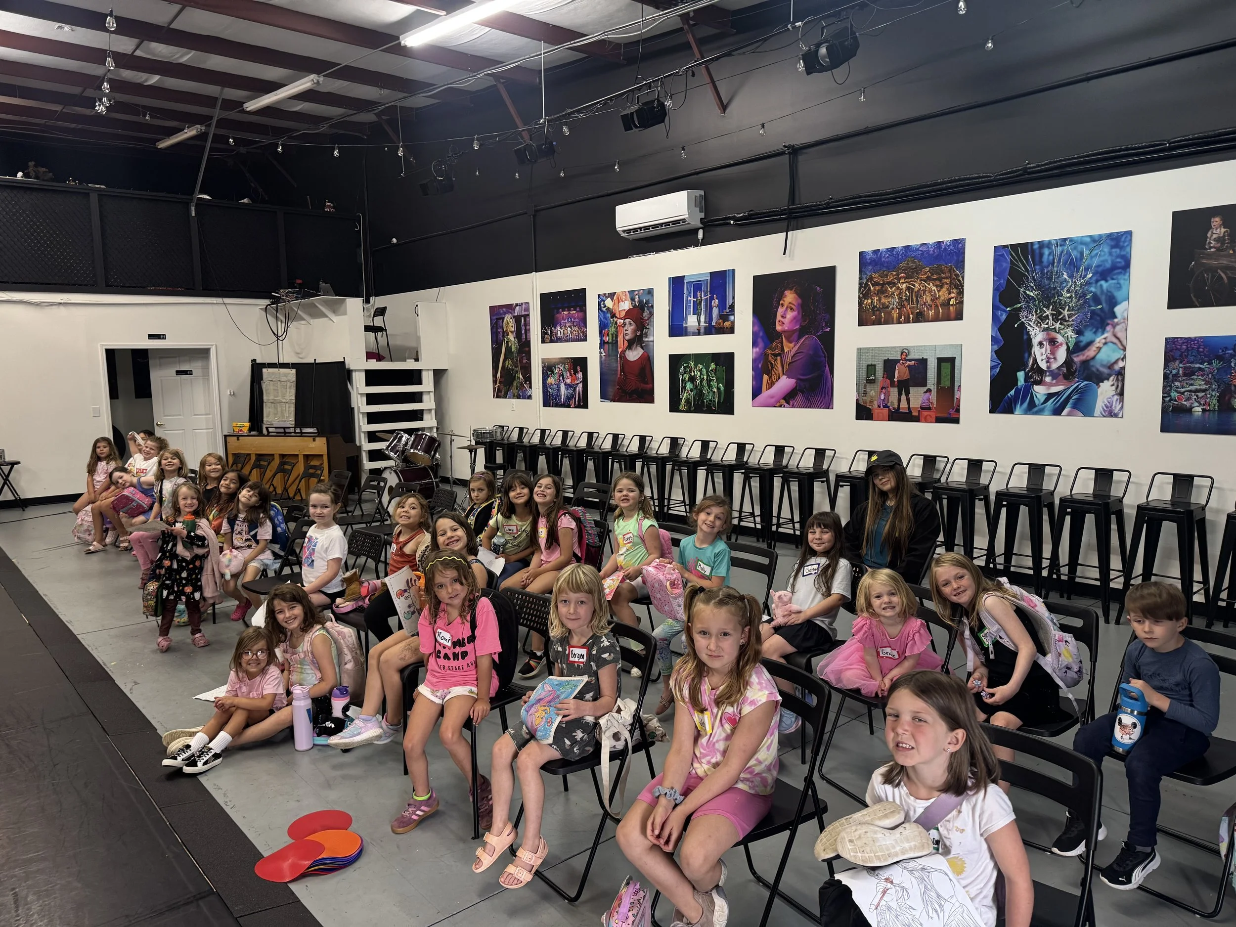 Children sitting and smiling on chairs in an indoor theater or classroom, with colorful posters on the wall behind them.