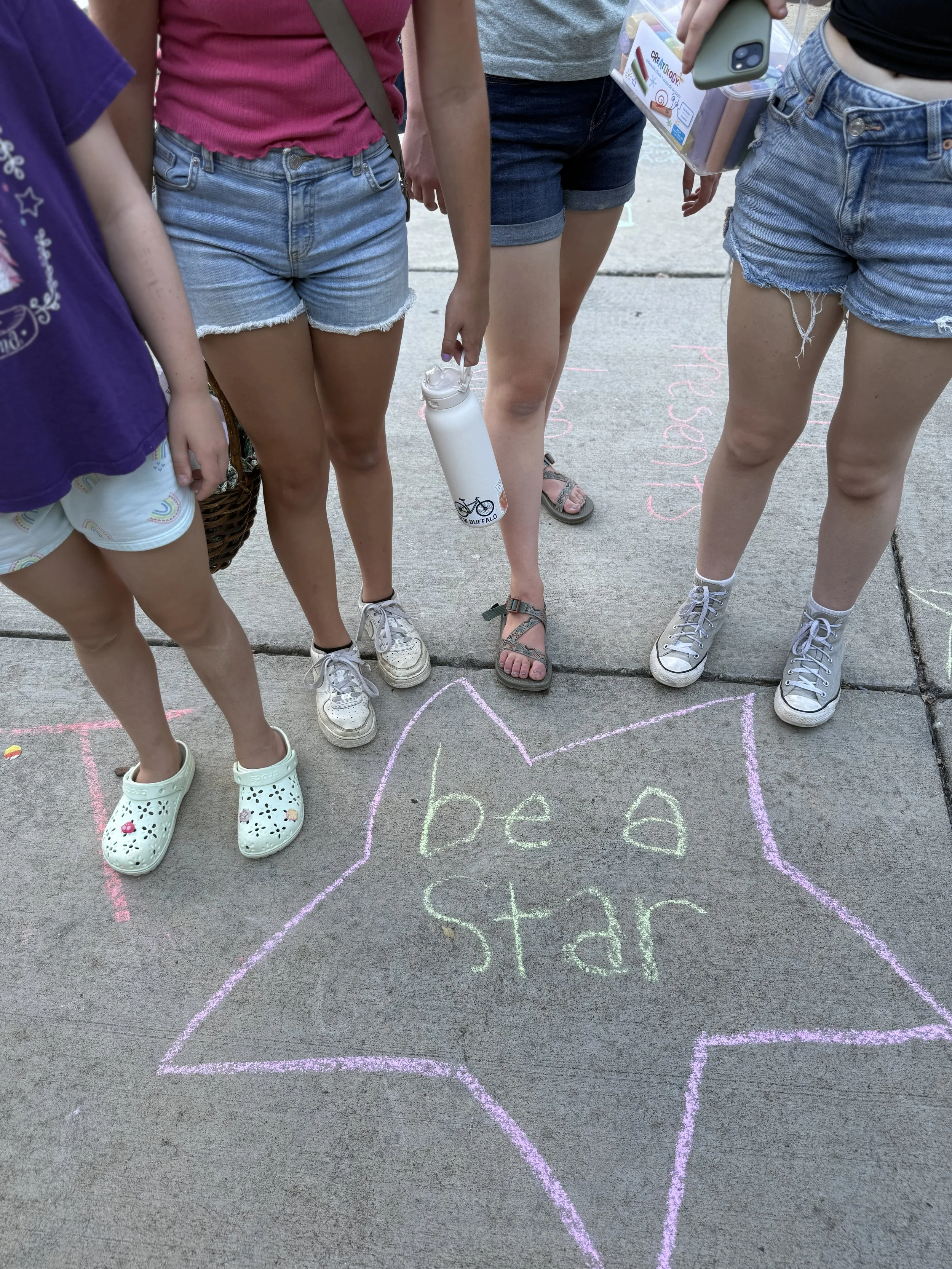 People standing around a sidewalk chalk drawing of a star with the words 'be a star' inscribed inside. Only their legs and part of their torsos are visible.
