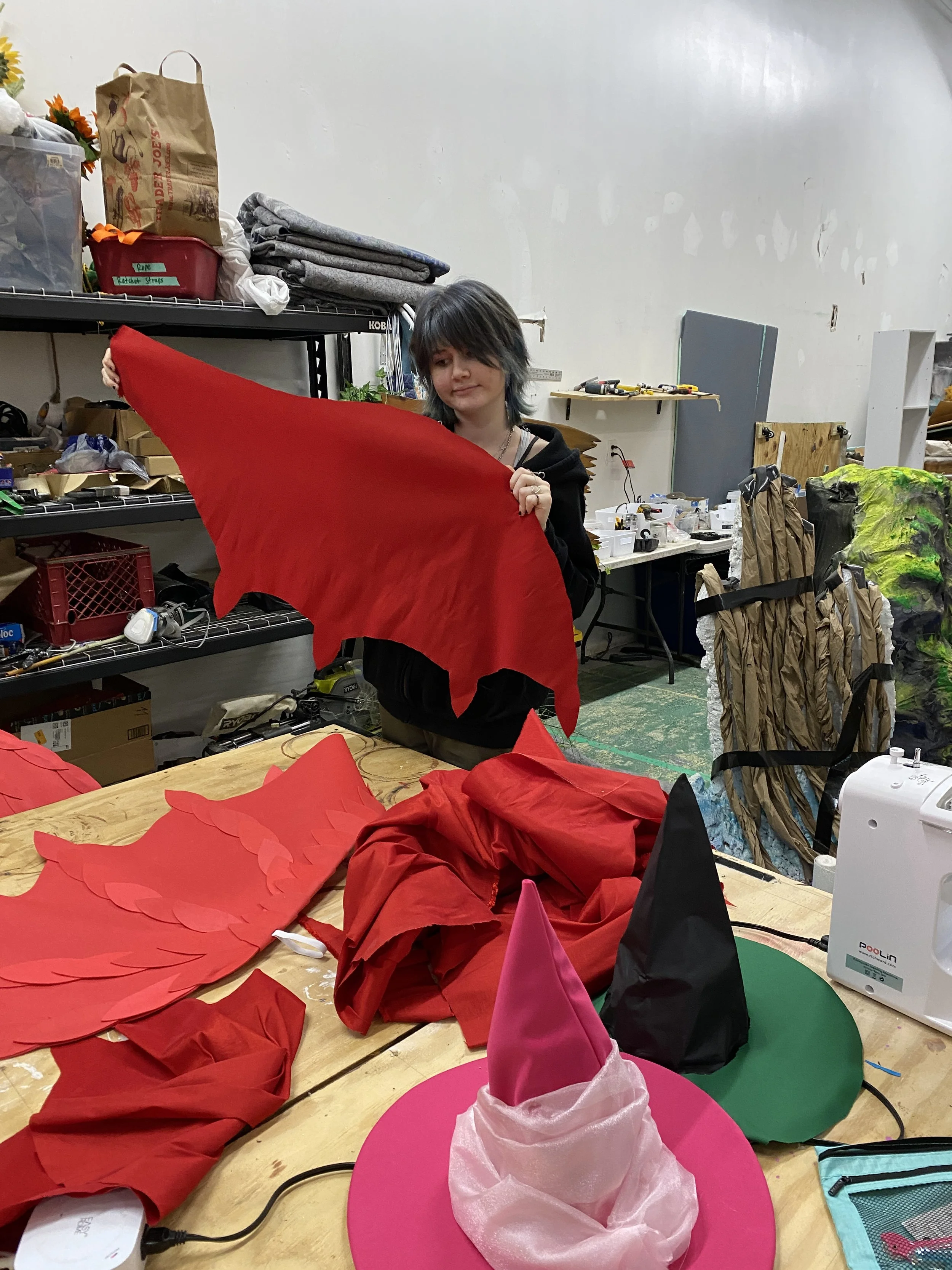 A person with teal and black hair working in a craft or costume workshop, holding a large red fabric piece. The table has red fabric, pink and black hats, and a pink hat with fabric. Shelves behind contain various crafting supplies and materials.