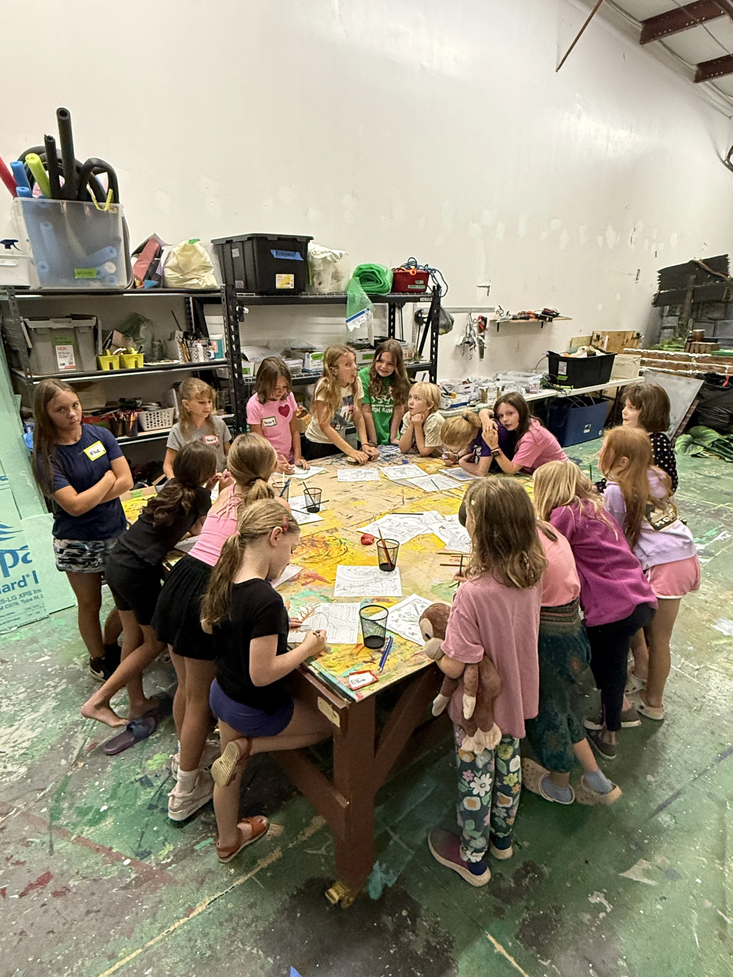 Group of young girls gather around a table in a workshop or art room, working on craft projects with papers, coloring supplies, and a stuffed animal, with shelves and supplies in the background.