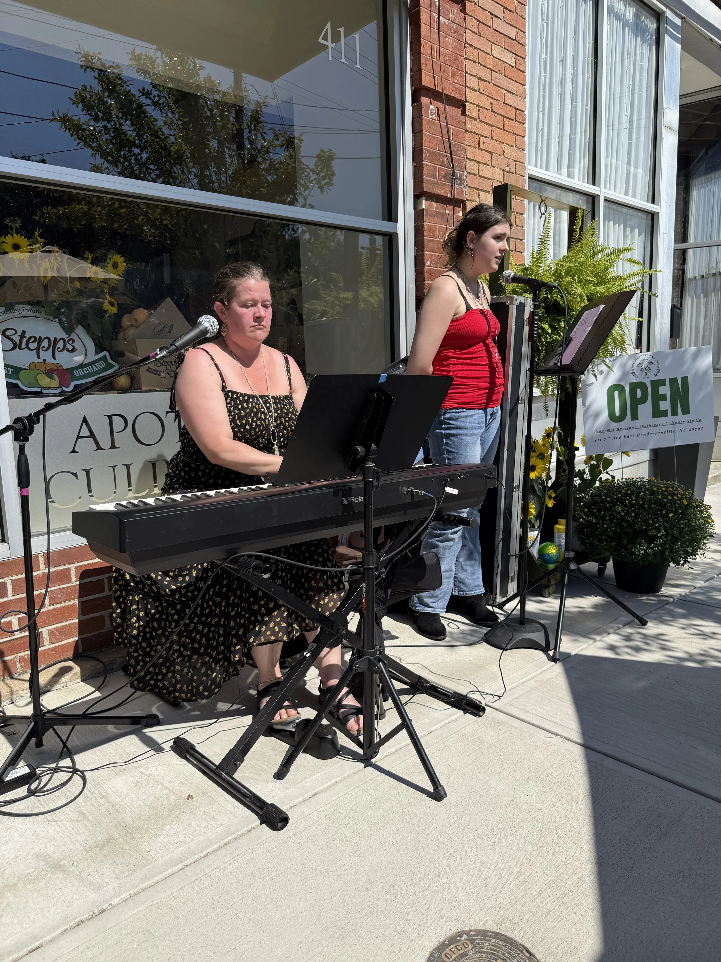 Two women performing music outside a storefront. One woman is playing a keyboard and singing, while the other stands and sings into a microphone. The storefront has large windows, brick accents, and an "OPEN" sign, with potted plants and flowers near