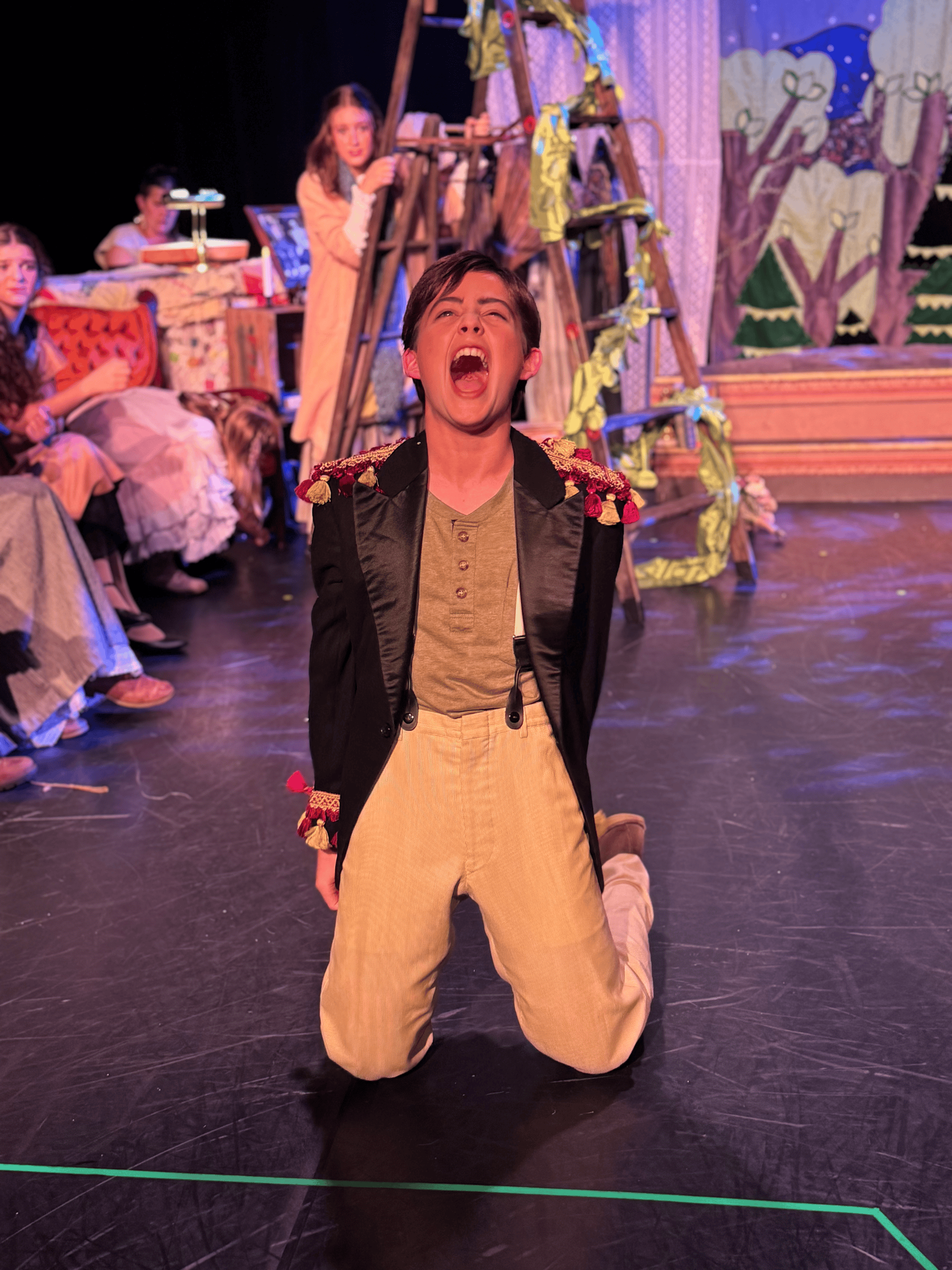 A young man is kneeling on the stage of a theater, singing passionately with his mouth wide open. He is dressed in a black jacket with decorative trim on the shoulders, a tan shirt, and beige pants. Behind him, a set with painted trees and a ladder i
