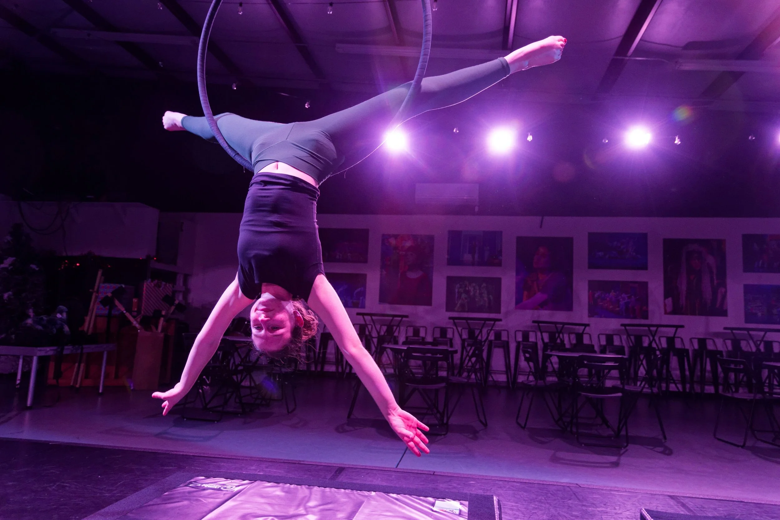 A woman performing an aerial acrobatics act on a hoop, hanging upside down in a dark room illuminated with purple lights.