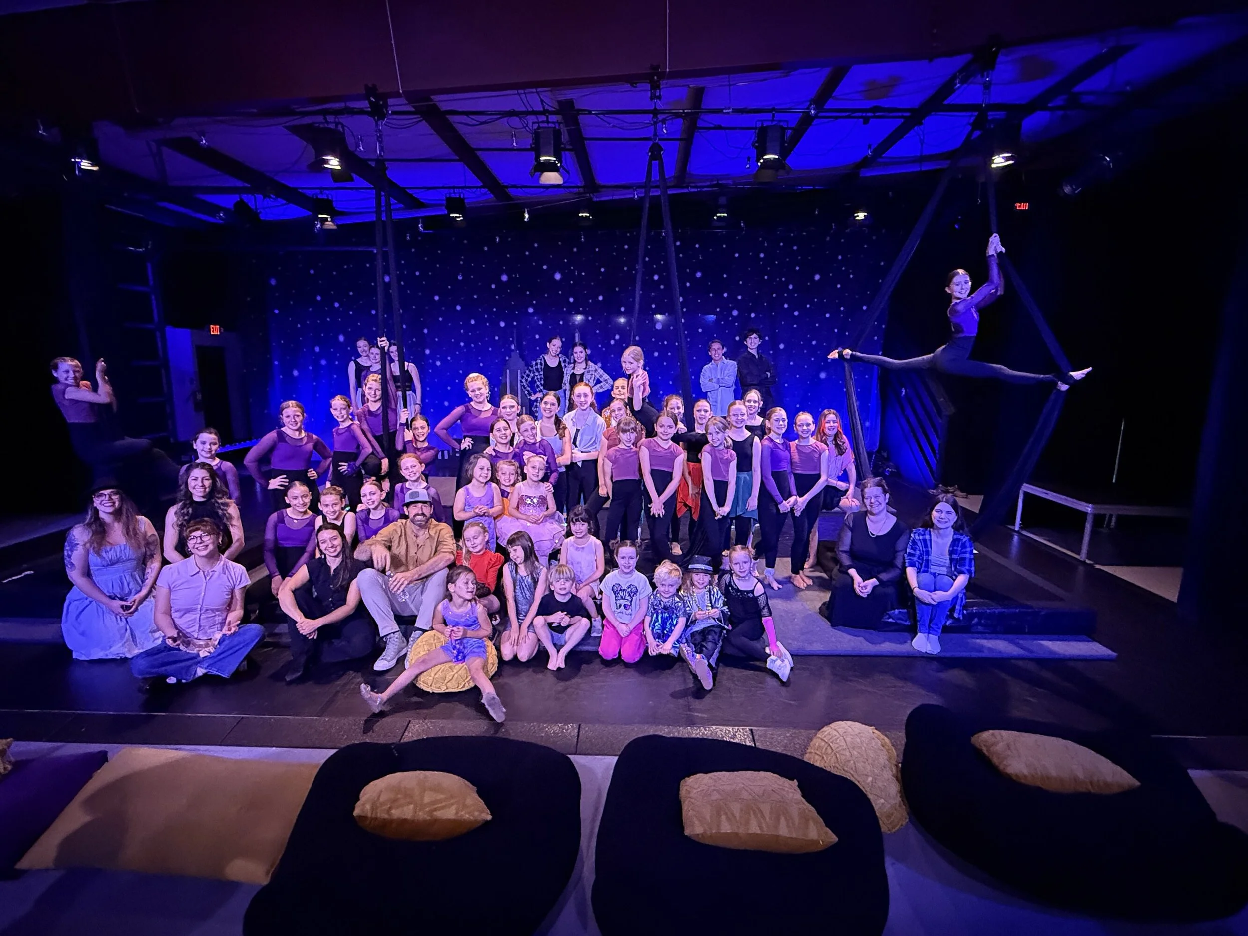 Group of young performers on stage in a theater, with some in costumes and others in casual clothing, after a dance or acrobatic performance, with a starry backdrop and aerial silks.