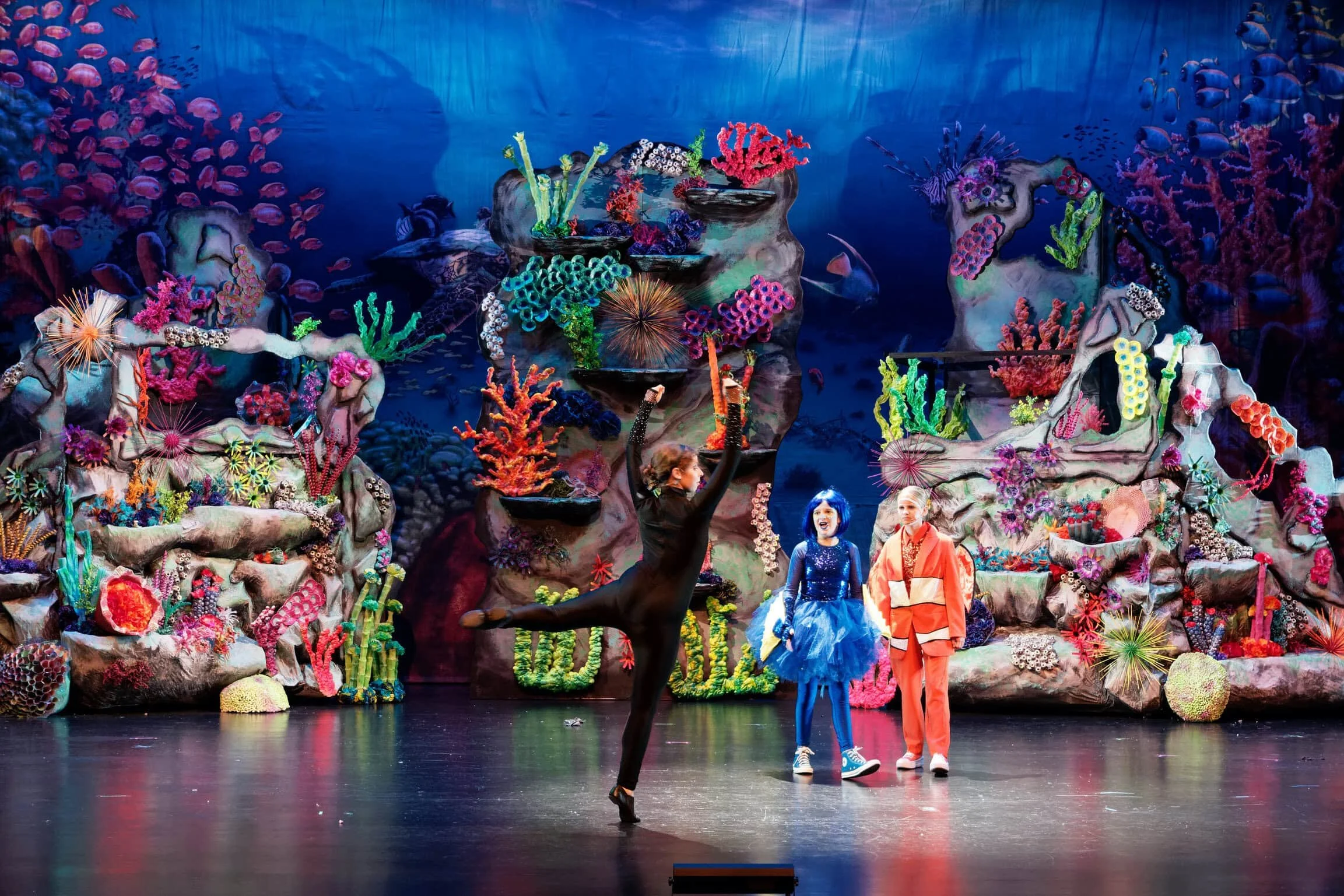 The image shows a stage with an underwater coral reef backdrop and two children dressed in costumes, one as a mermaid and another as a character in an orange outfit. A performer in black is dancing with one leg extended horizontally.