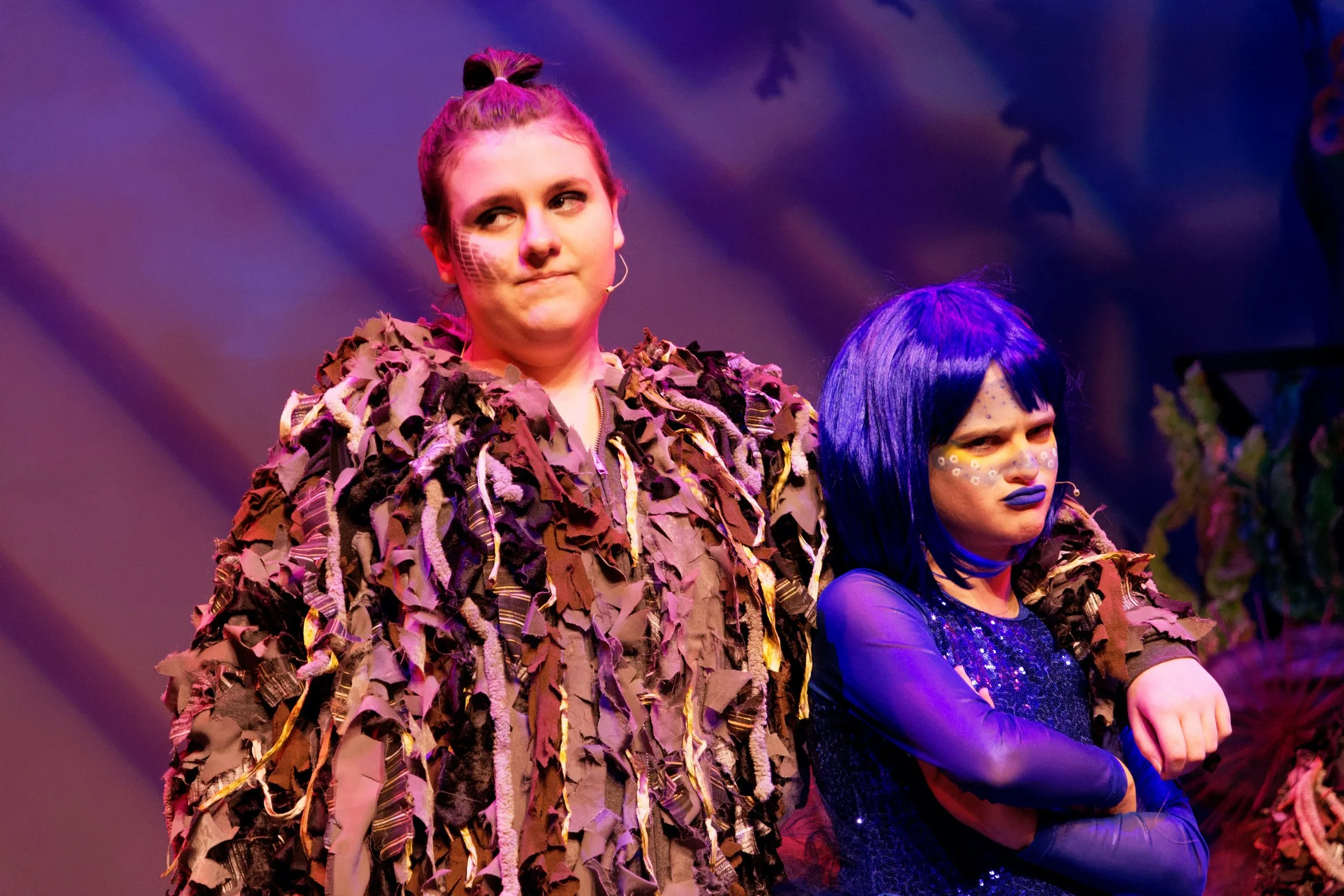 Two women in theatrical costumes, one with a leaf-covered jacket and the other with blue hair and makeup, posing on stage.