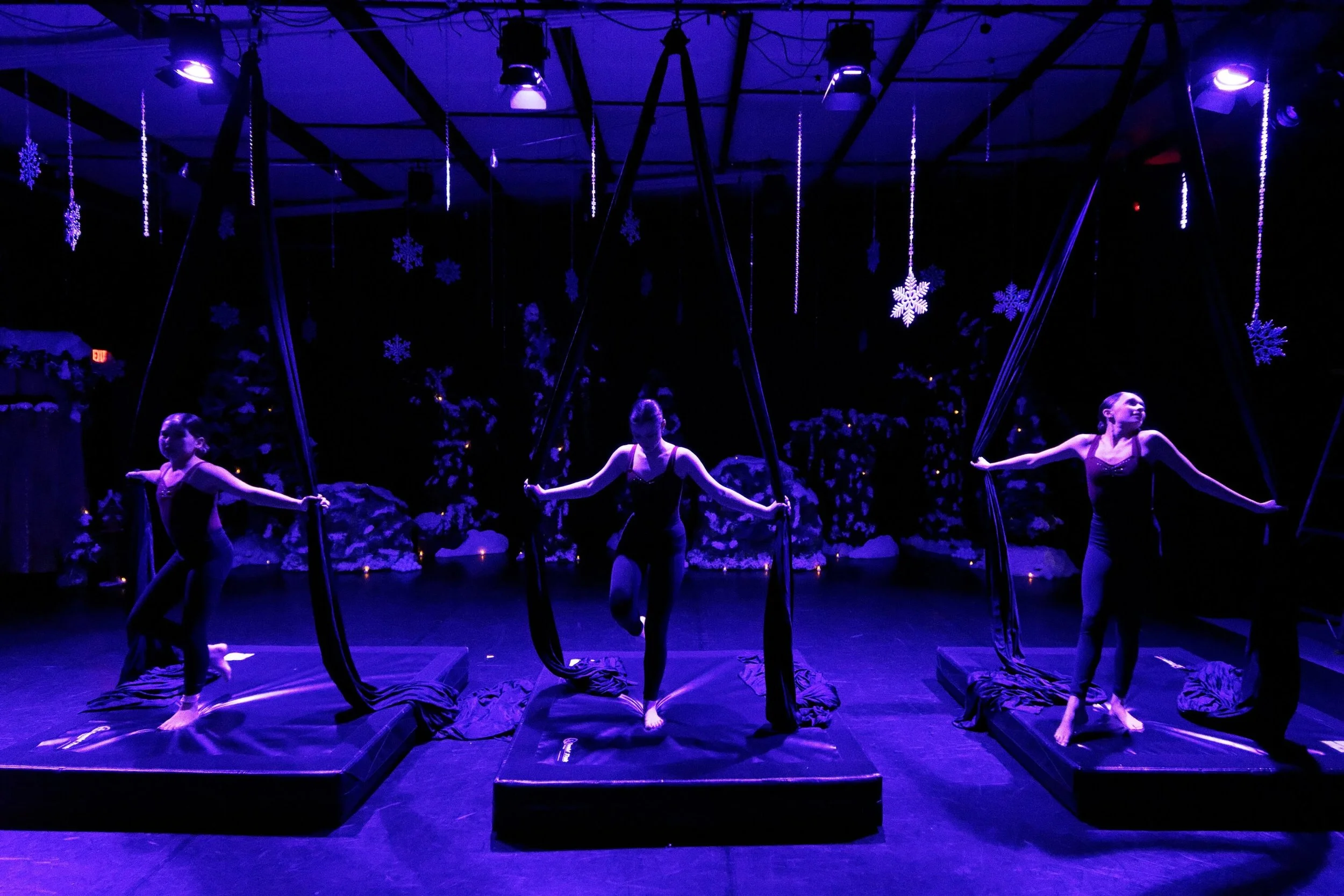Three female aerial silk performers practicing on stage under blue purple lighting with snowflake decorations hanging from the ceiling and snow-covered trees in the background.
