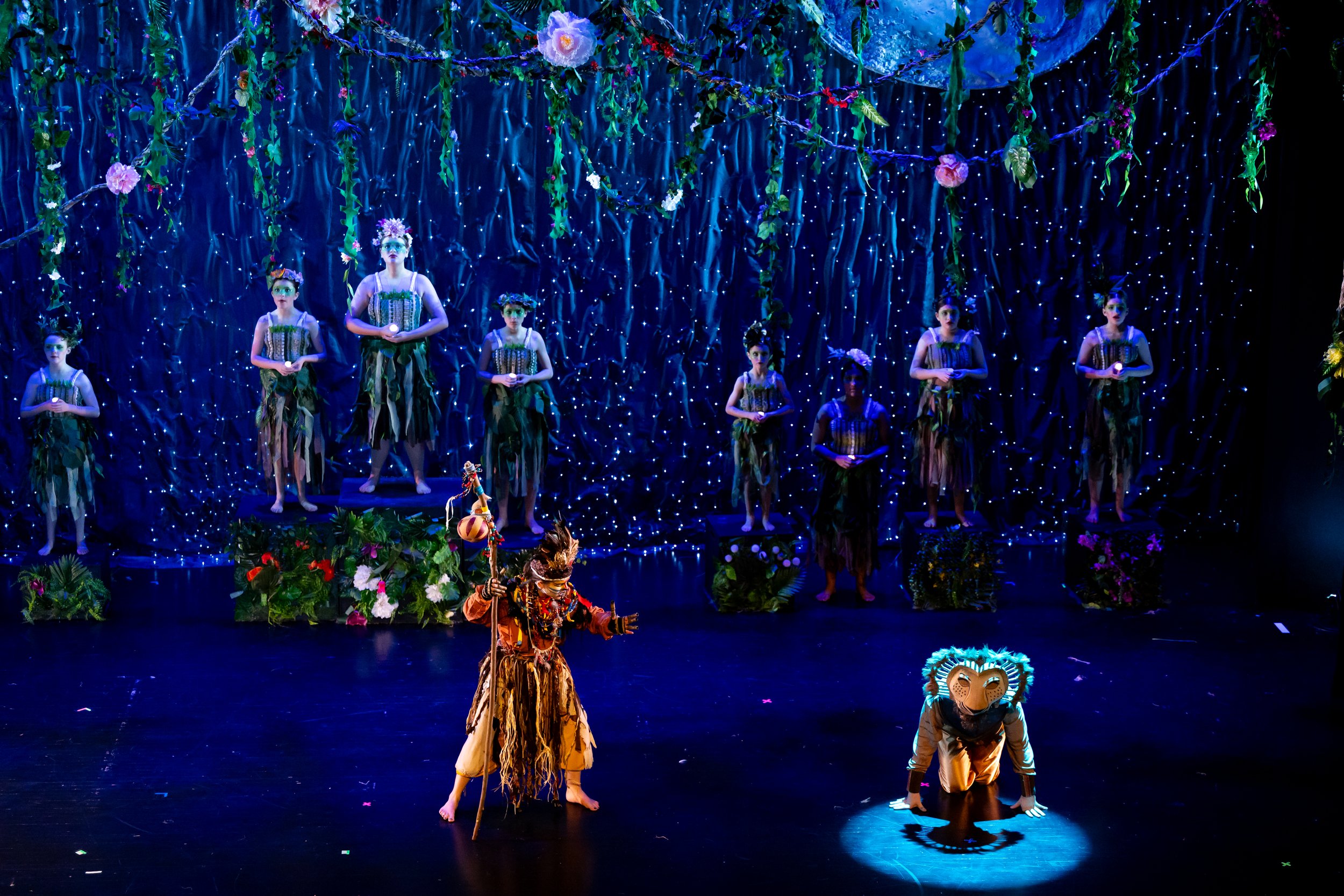 The stage features a theatrical performance inspired by 'The Lion King JR,' with actors in animal costumes, including lions and jungle characters, against a vibrant jungle-themed backdrop with blue lighting, flowers, and hanging vines. Simba, Nala, R