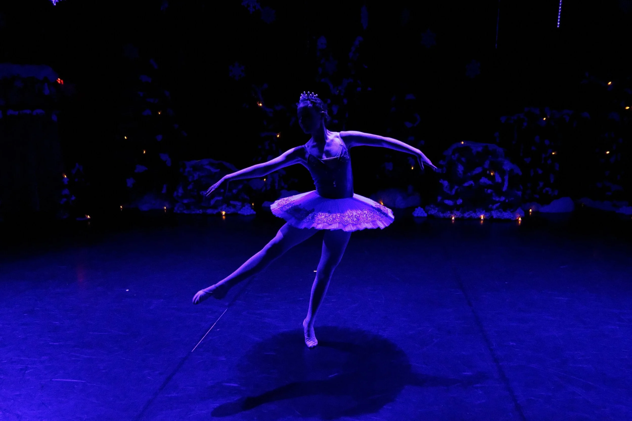 A ballet dancer performing on stage under purple lighting, wearing a tutu and a tiara, with background decorated with snowflakes and small lights.