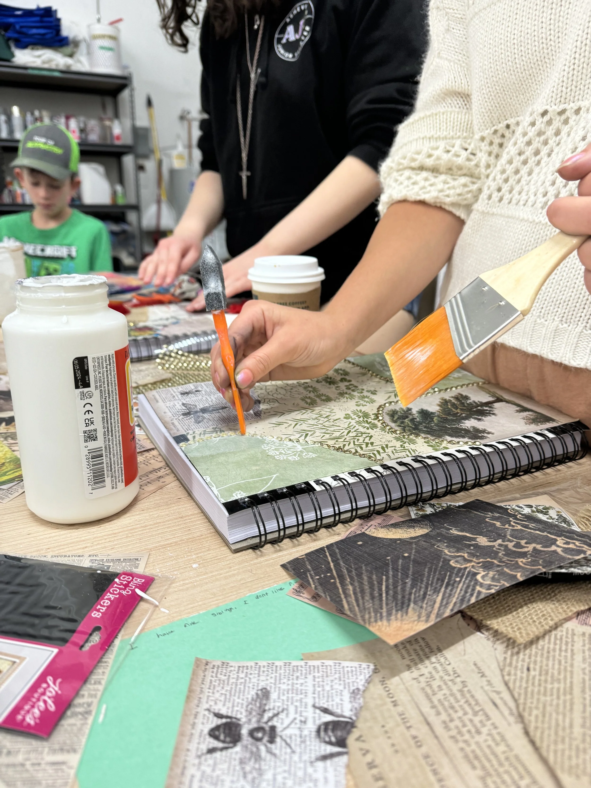 People working on a craft project at a table, with art supplies including paintbrushes, glue, and decorative paper, in a workshop or classroom setting.