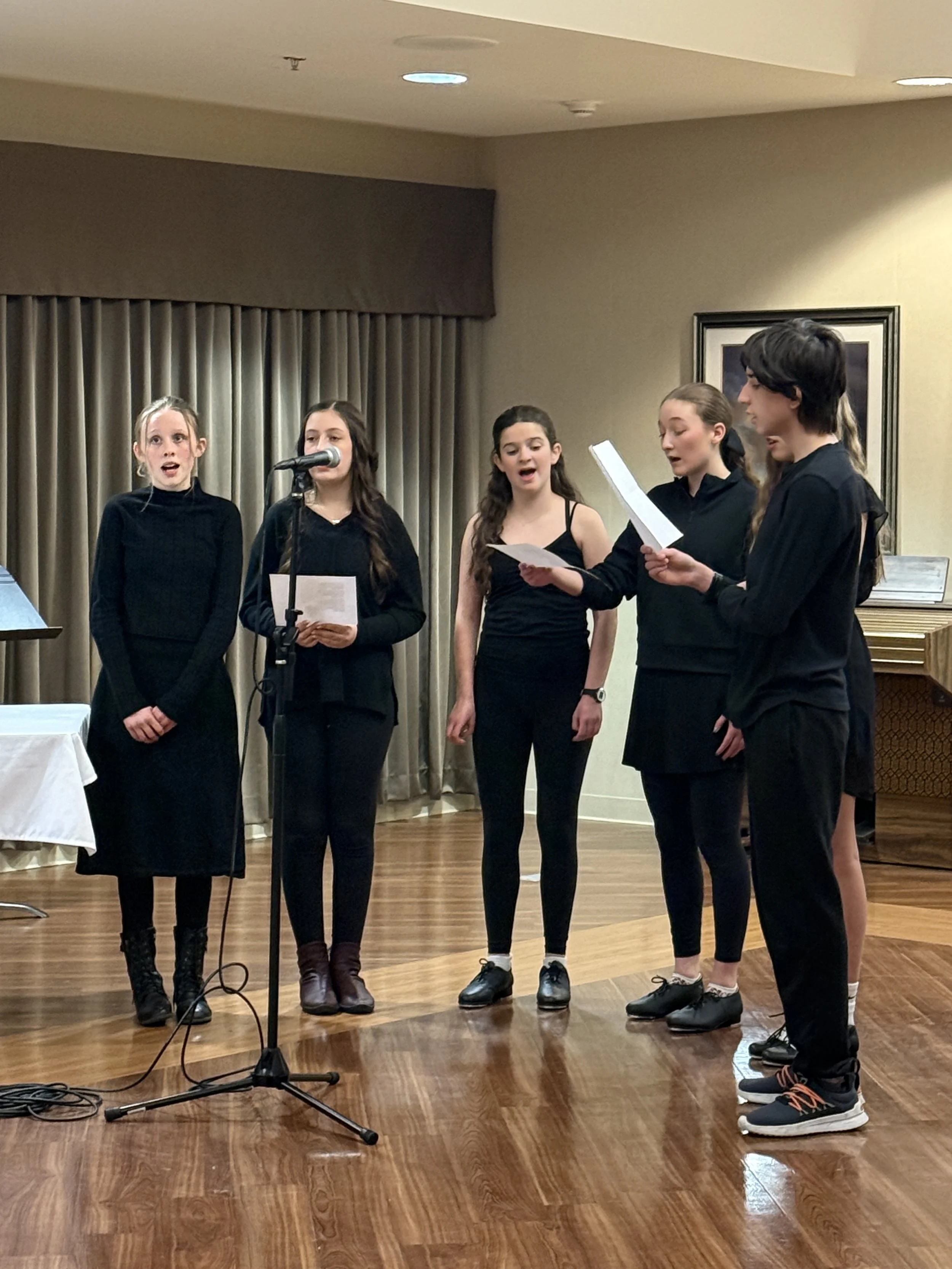 Five young girls standing on a wooden floor, singing or speaking into a microphone and reading from papers, in a room with beige walls, closed curtains, and a framed picture.