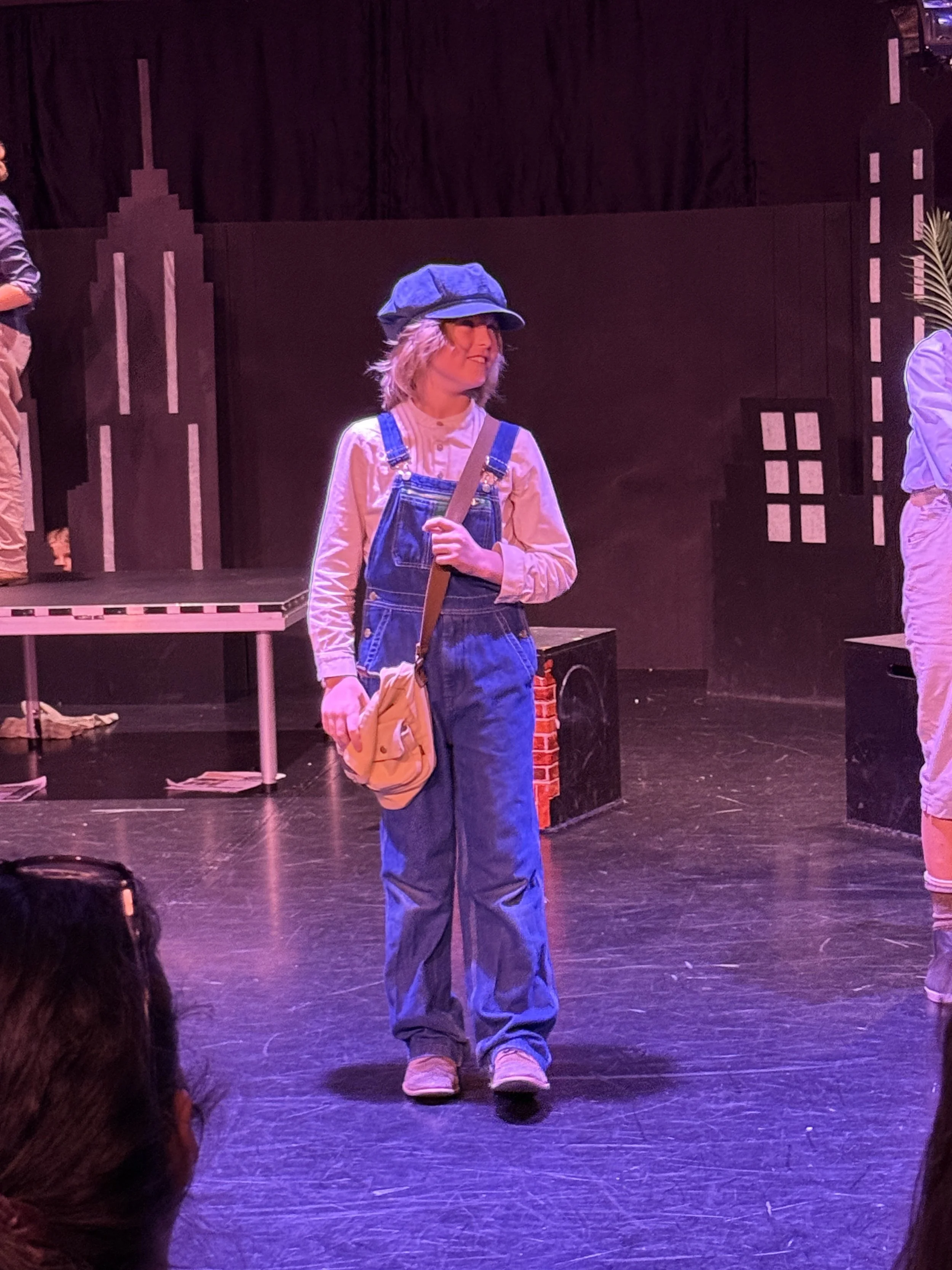 A woman on stage dressed in overalls, a long-sleeve shirt, and a cap, with a small beige bag across her body, in a theatrical setting with black walls and props.