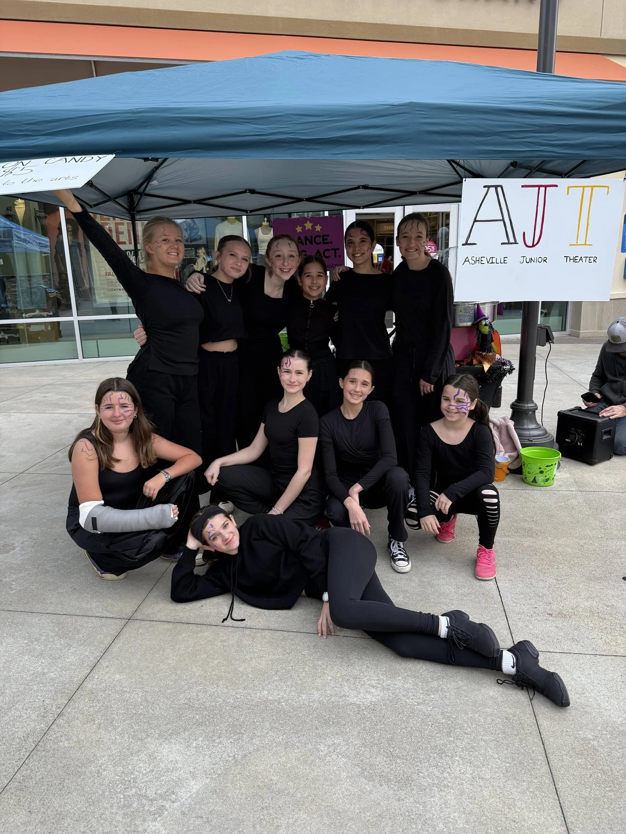 Girls in black costumes with face paint posing under a blue canopy at a junior theater event, with a sign reading 'AJT Asheville Junior Theater' in the background.
