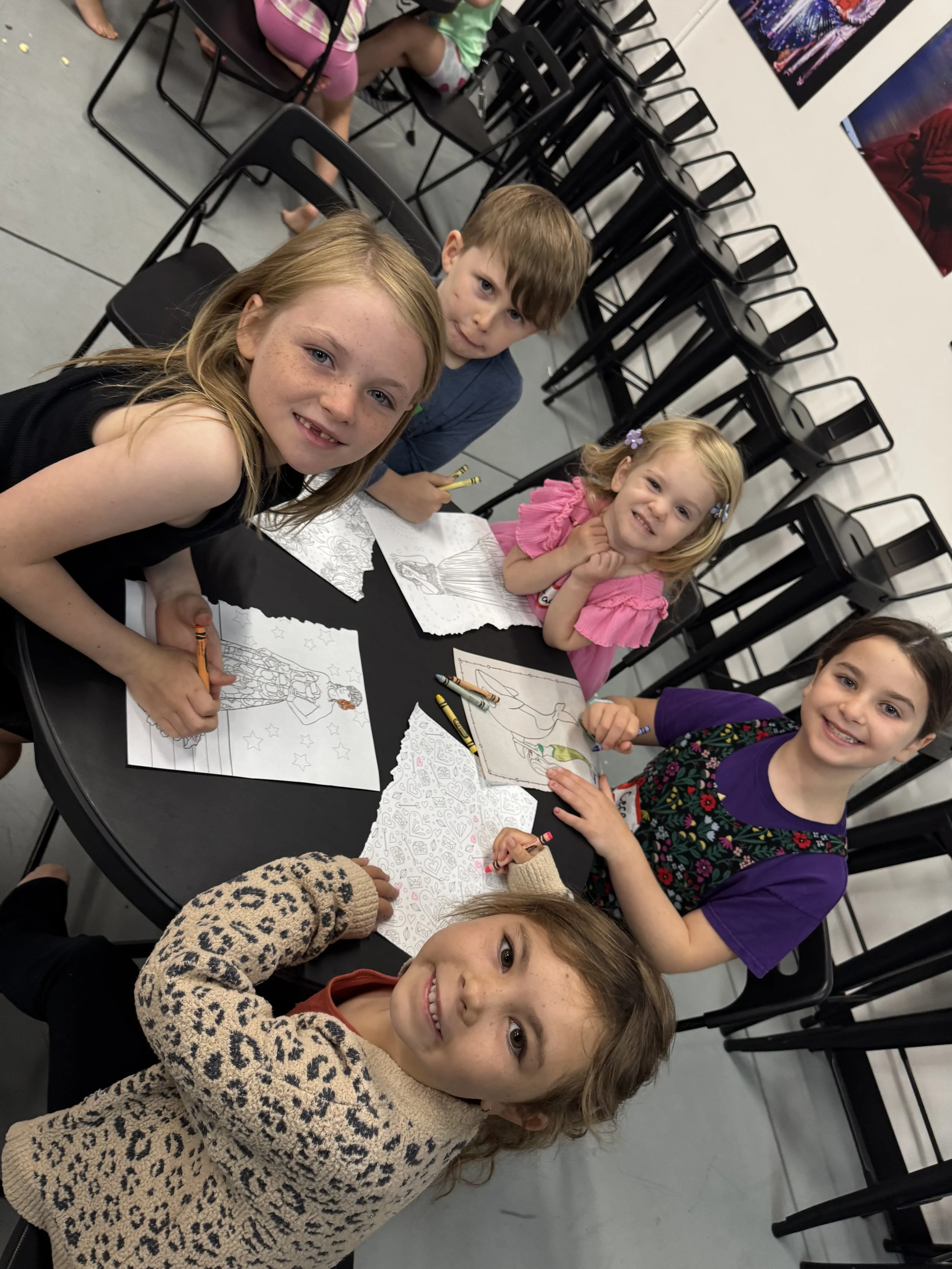 Six children are sitting around a black round table, coloring pictures on paper with crayons. The children are smiling and appear to be enjoying their activity in a room with stacked black chairs and colorful artwork on the walls.
