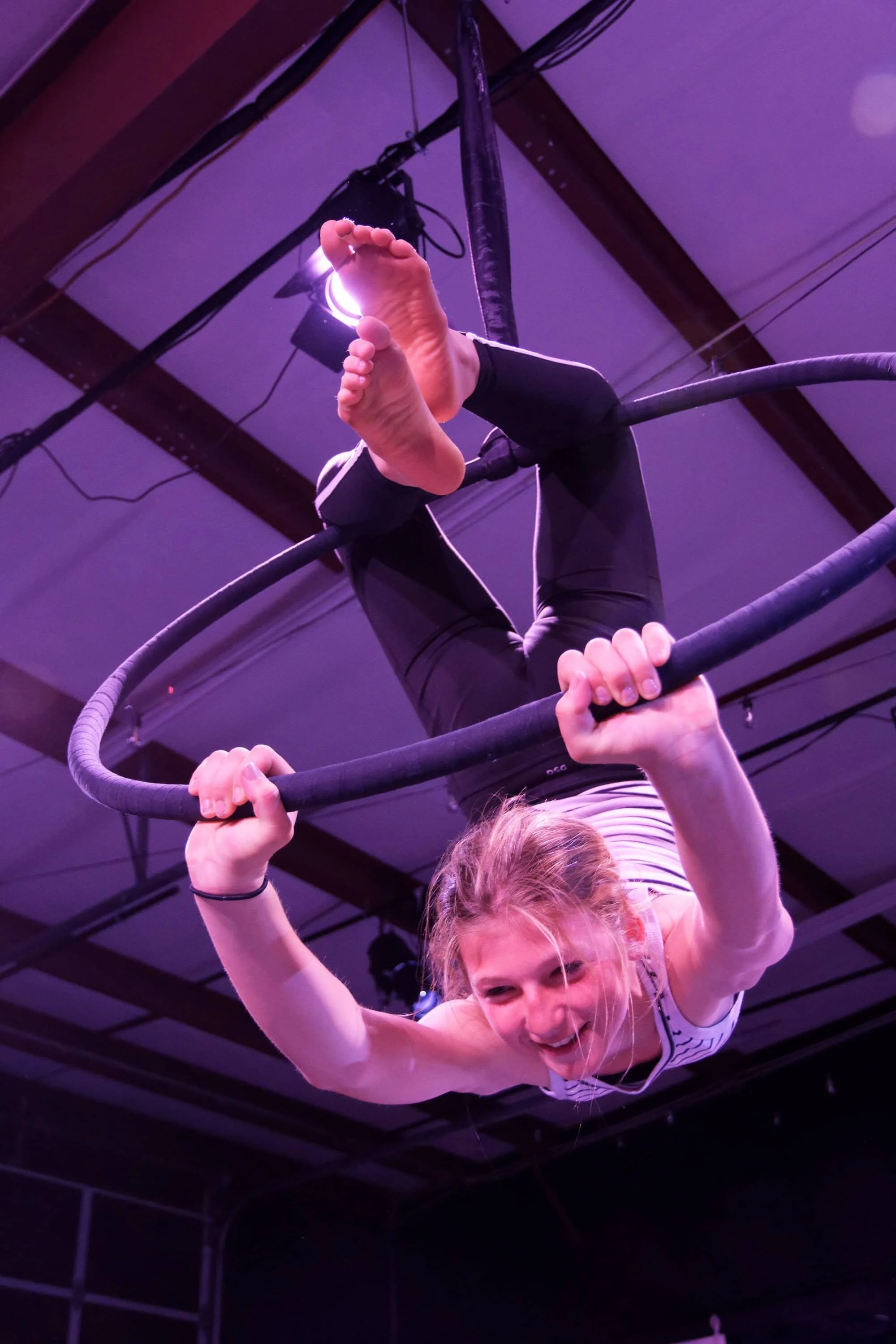 A young woman performing a trapeze act, hanging upside down from a trapeze bar with a big smile, in a venue illuminated with purple lighting.