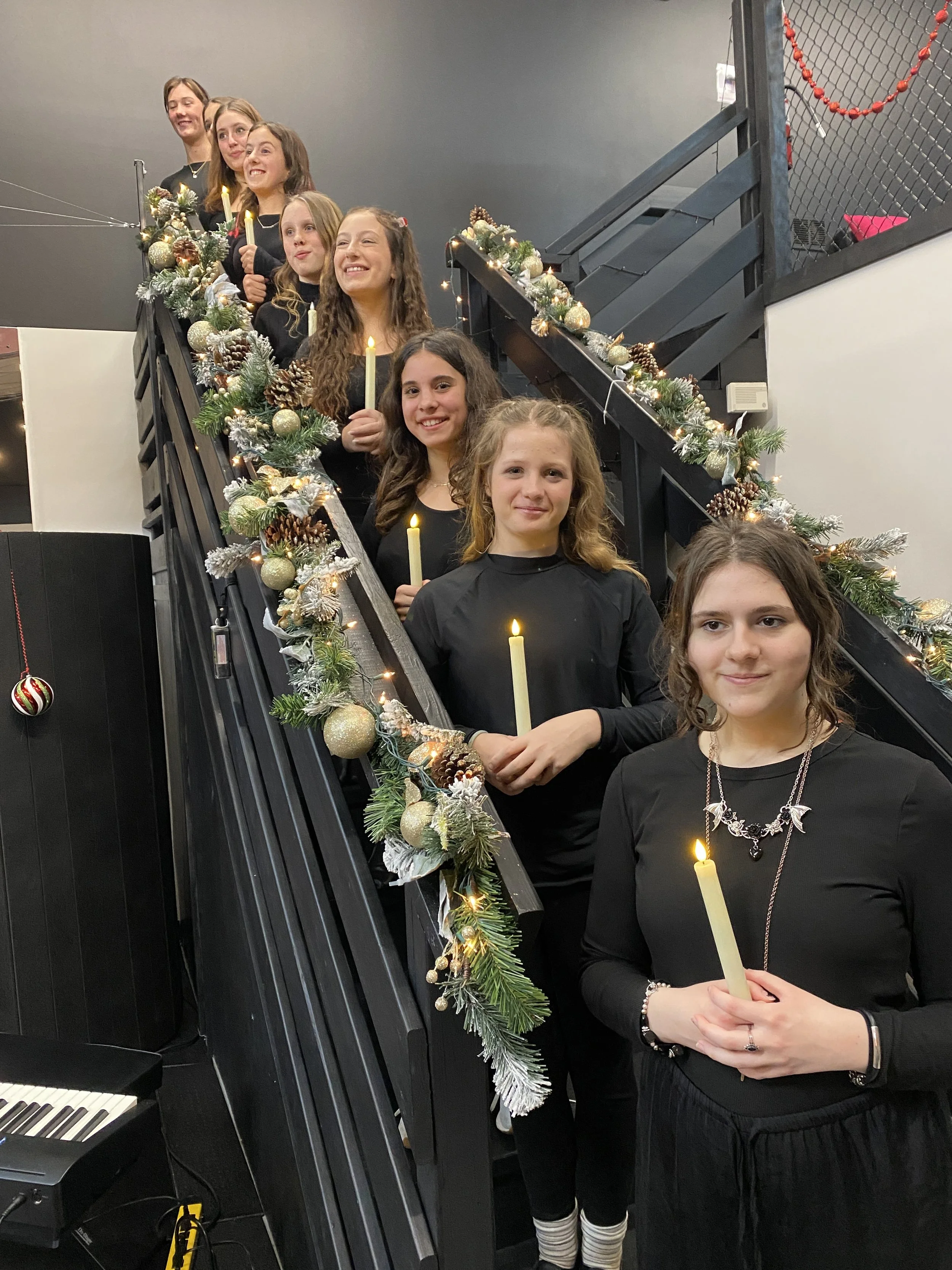 Group of nine young women standing on a decorated staircase holding candles, dressed in black for a holiday celebration.