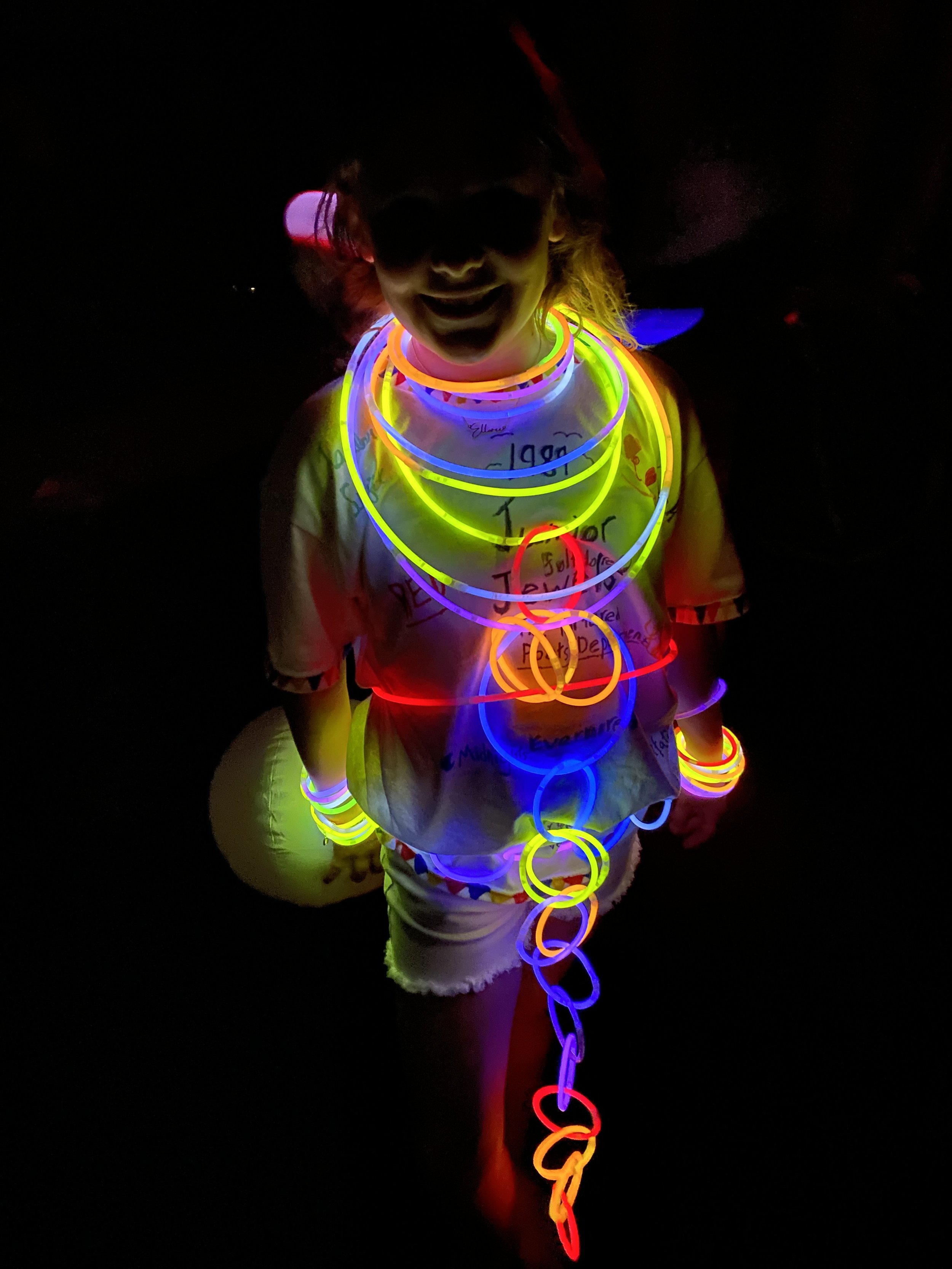 Person smiling in a dark environment, wearing multiple colorful glow-in-the-dark necklaces and bracelets, with some written text on their shirt.