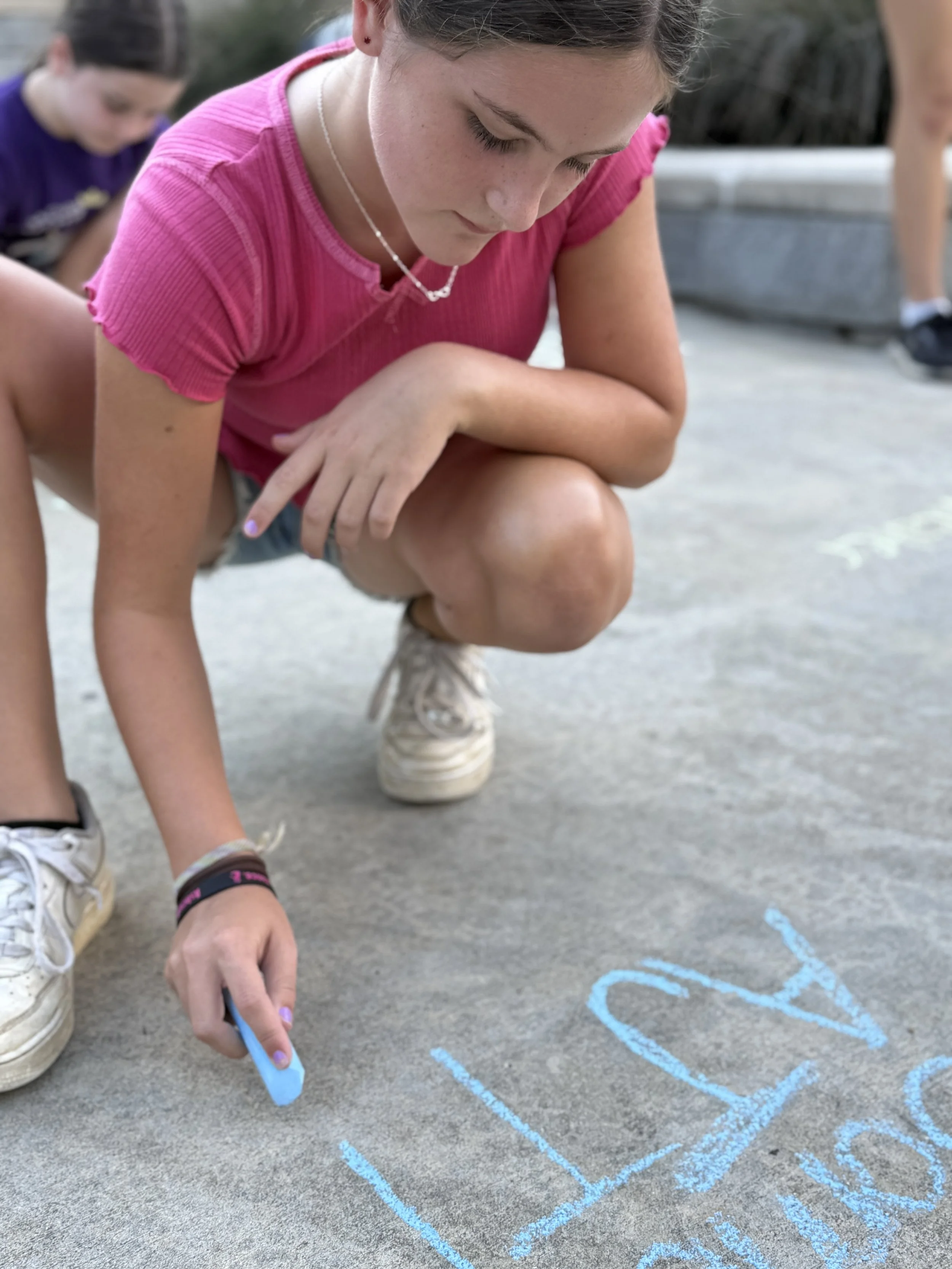 A girl in a pink shirt and shorts crouches on a concrete surface, writing with blue chalk.