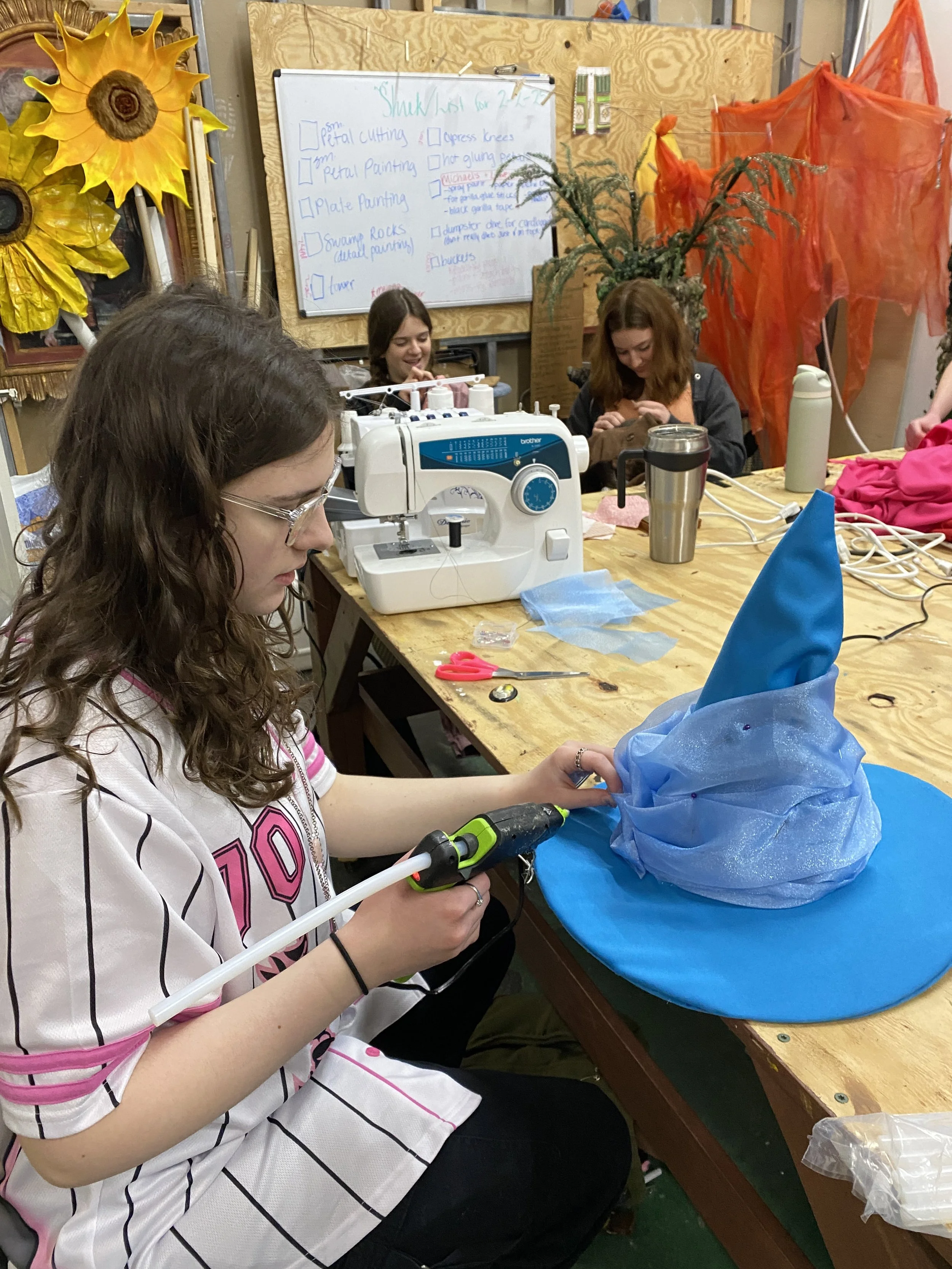 A girl using a hot glue gun to decorate a blue witch hat in a crafting workshop with other girls working at the table, surrounded by sewing supplies and Halloween decorations.