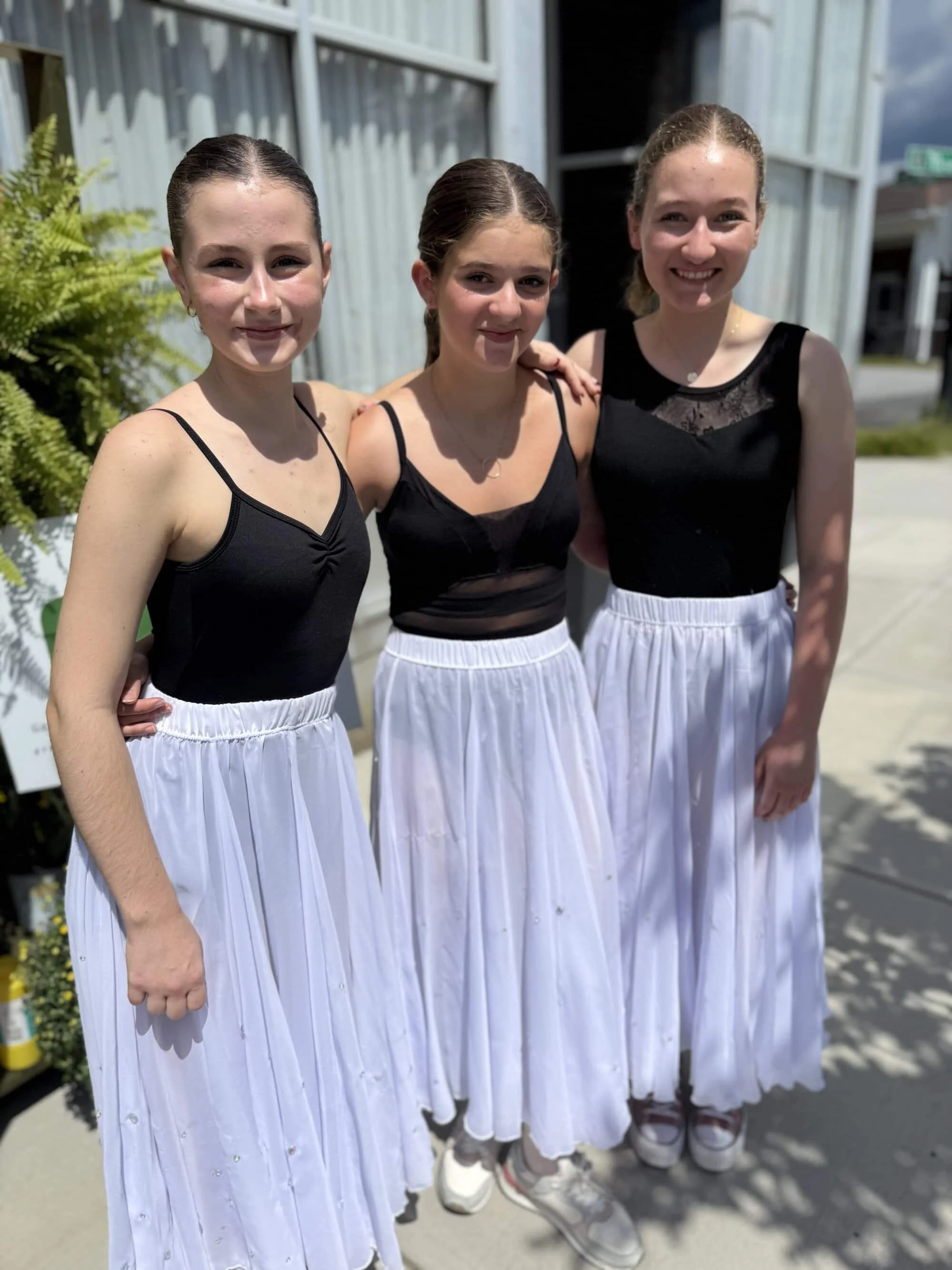 Three young girls standing outdoors in front of a building, dressed in black tank tops and white skirts, smiling at the camera.