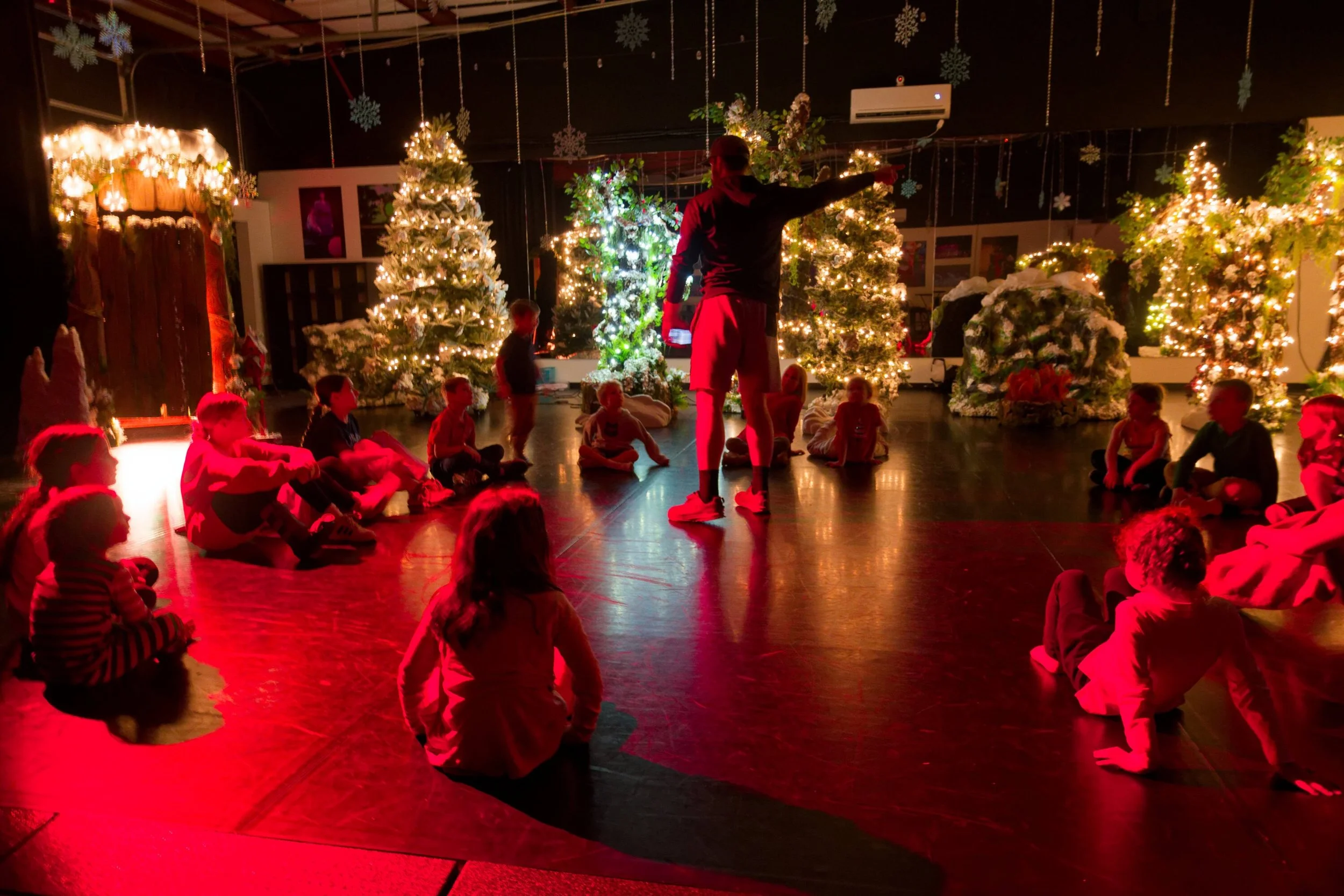 A group of children sitting on the floor in a circular formation, attentively listening to an adult presenter standing in the center. The room is decorated with Christmas trees adorned with lights, ornaments, and faux snow, as well as hanging snowfla