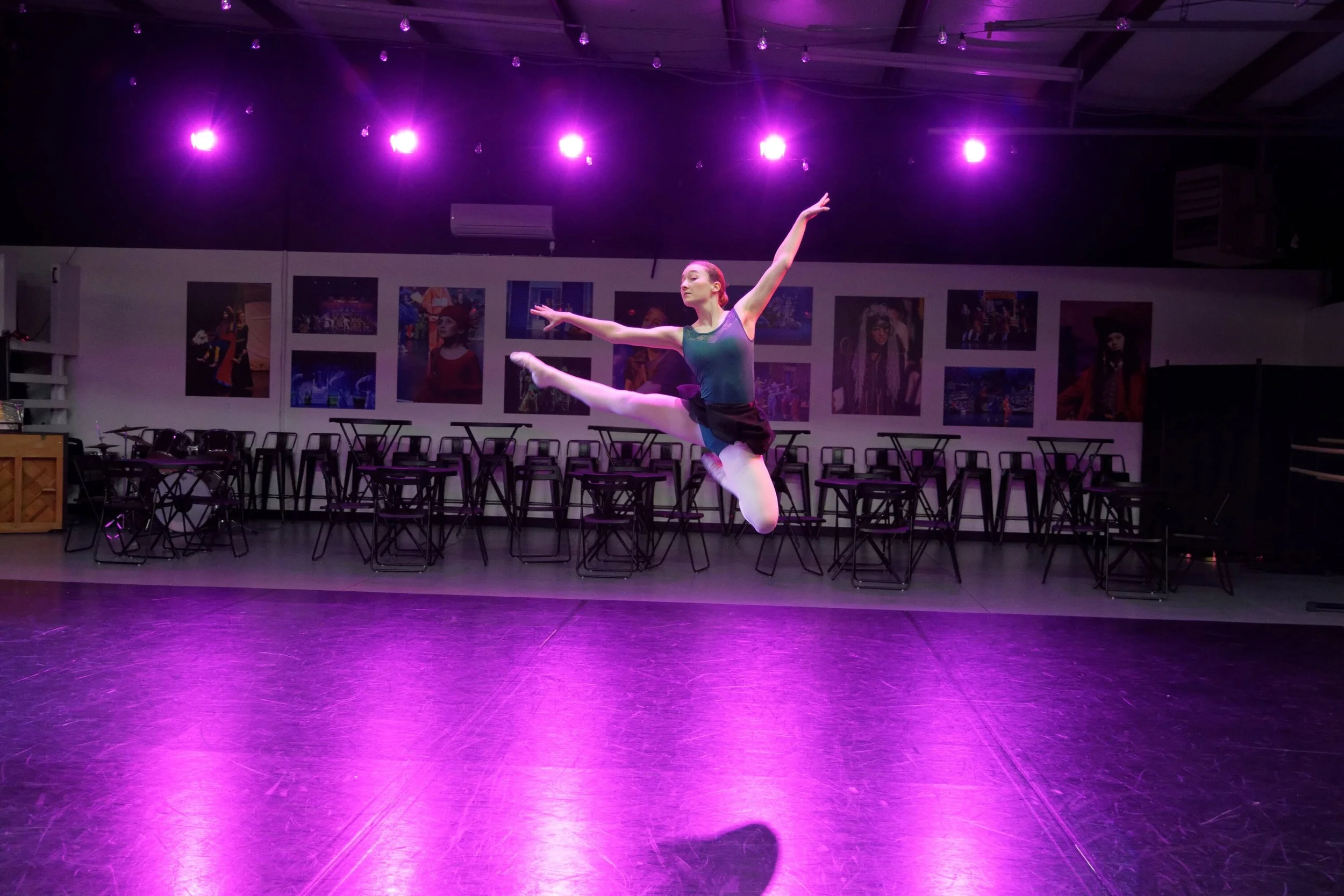 A ballet dancer mid-air leap in a dance studio with purple lighting, empty tables and chairs, and colorful artwork on the walls.