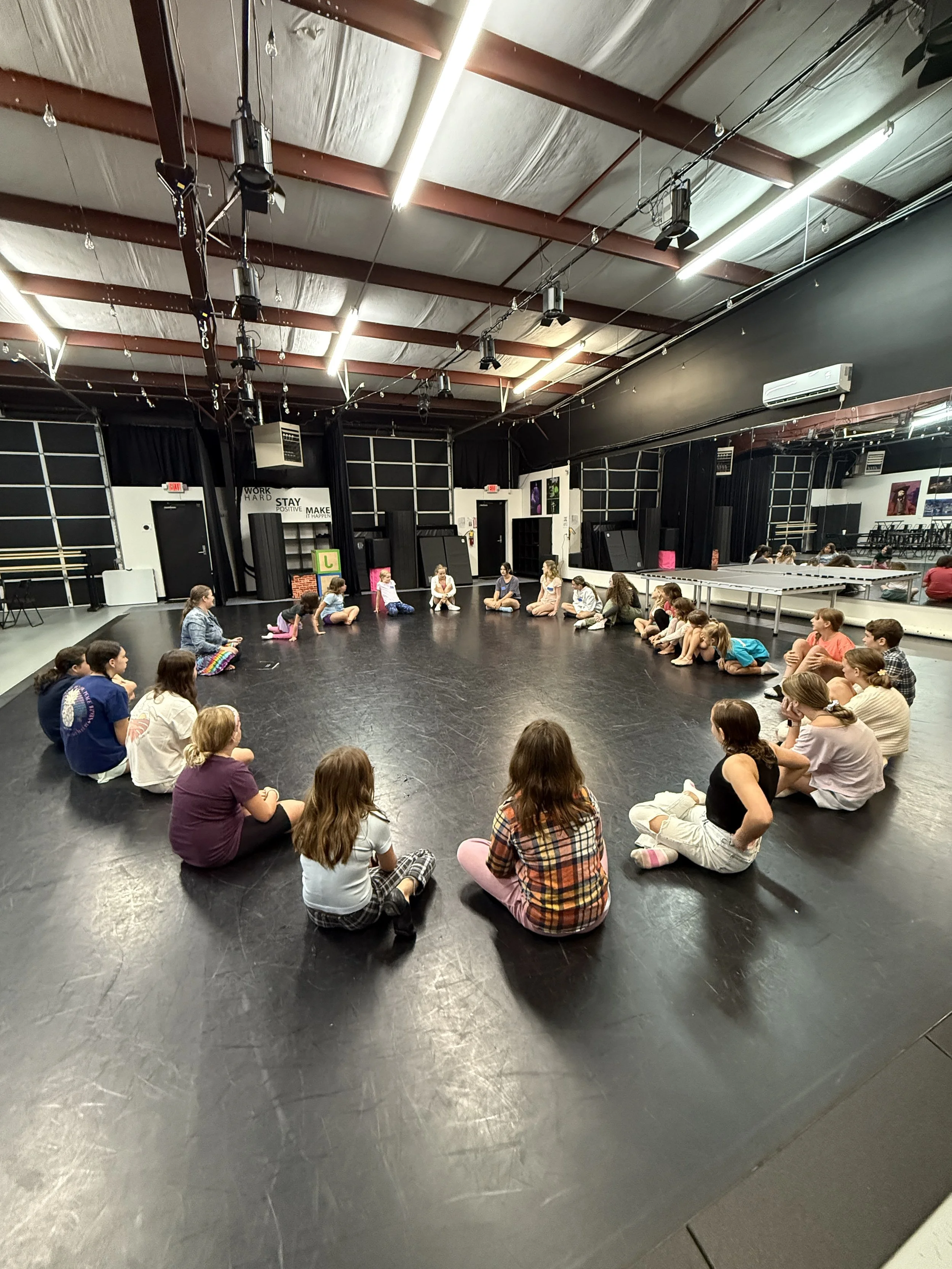 Children sitting in a circle in a dance studio with black flooring and high ceilings.