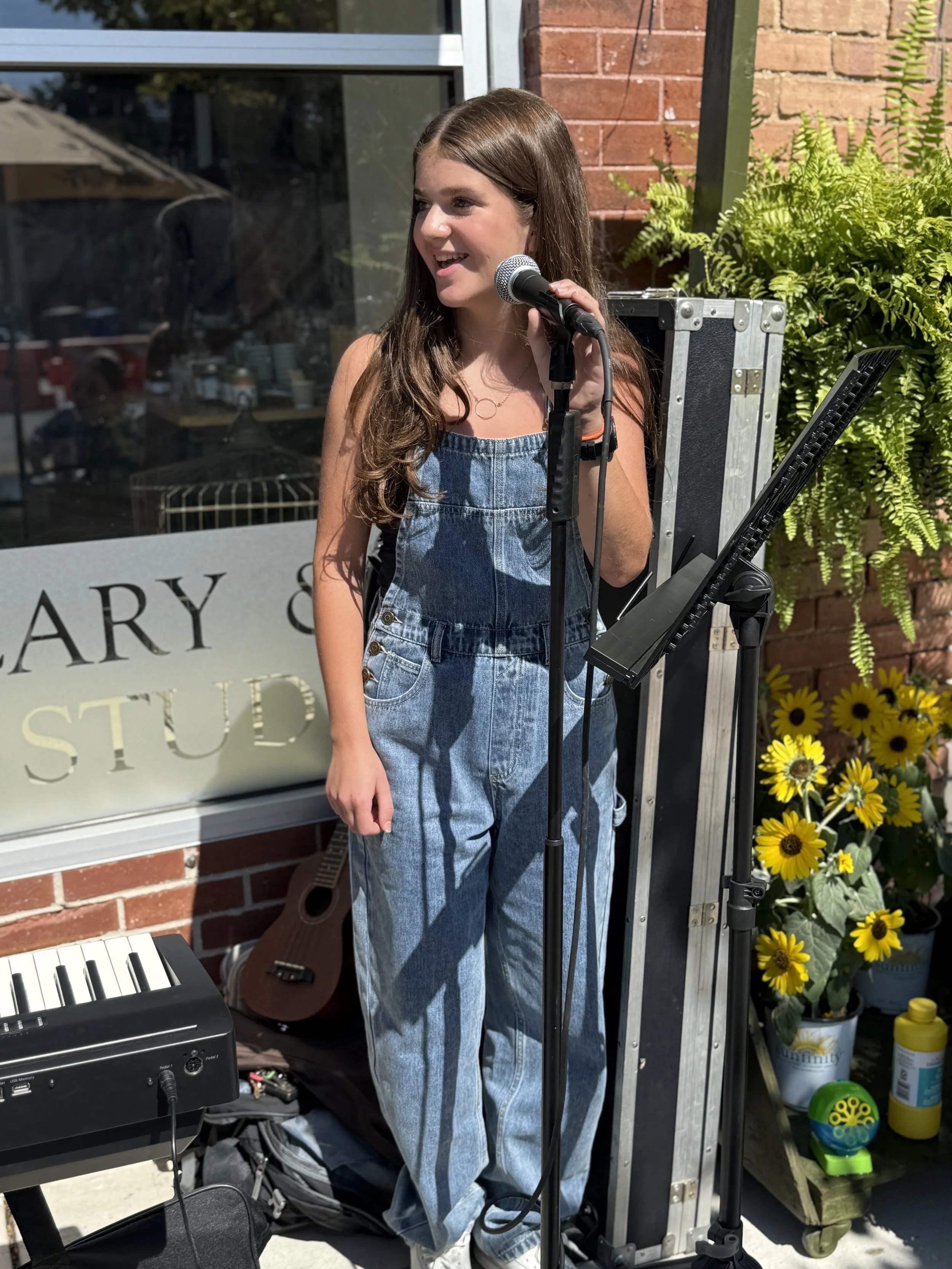 A young girl with long brown hair stands outside, holding a microphone in her hand, smiling, and appears to be singing or speaking. She is wearing denim overalls and is standing next to a music stand, with a keyboard and guitar nearby. Behind her, th