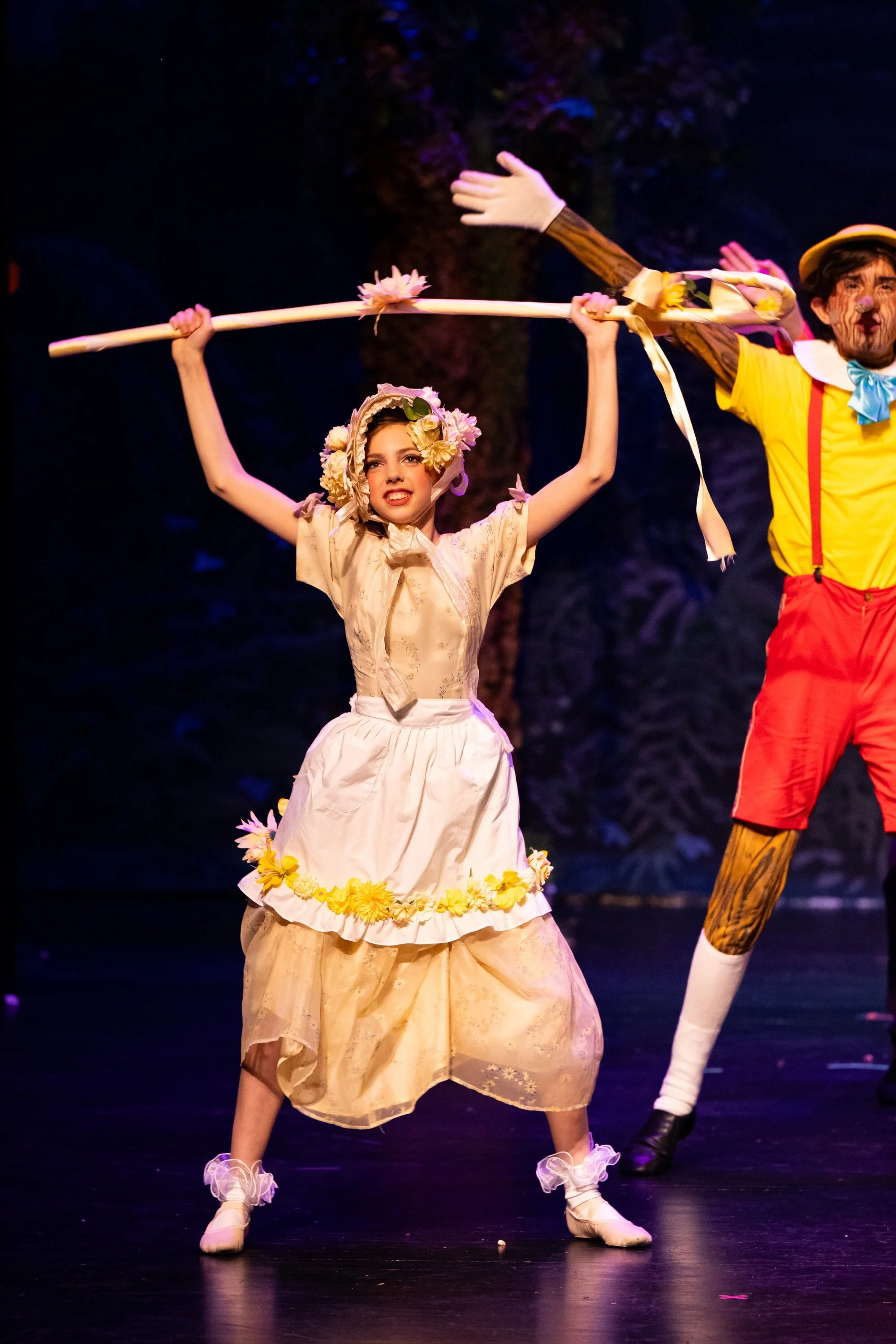 Two actors on stage dressed in costumes, with the woman in a vintage dress and floral headpiece holding a long stick with flowers, and the man in bright clothing, in a theatrical performance.