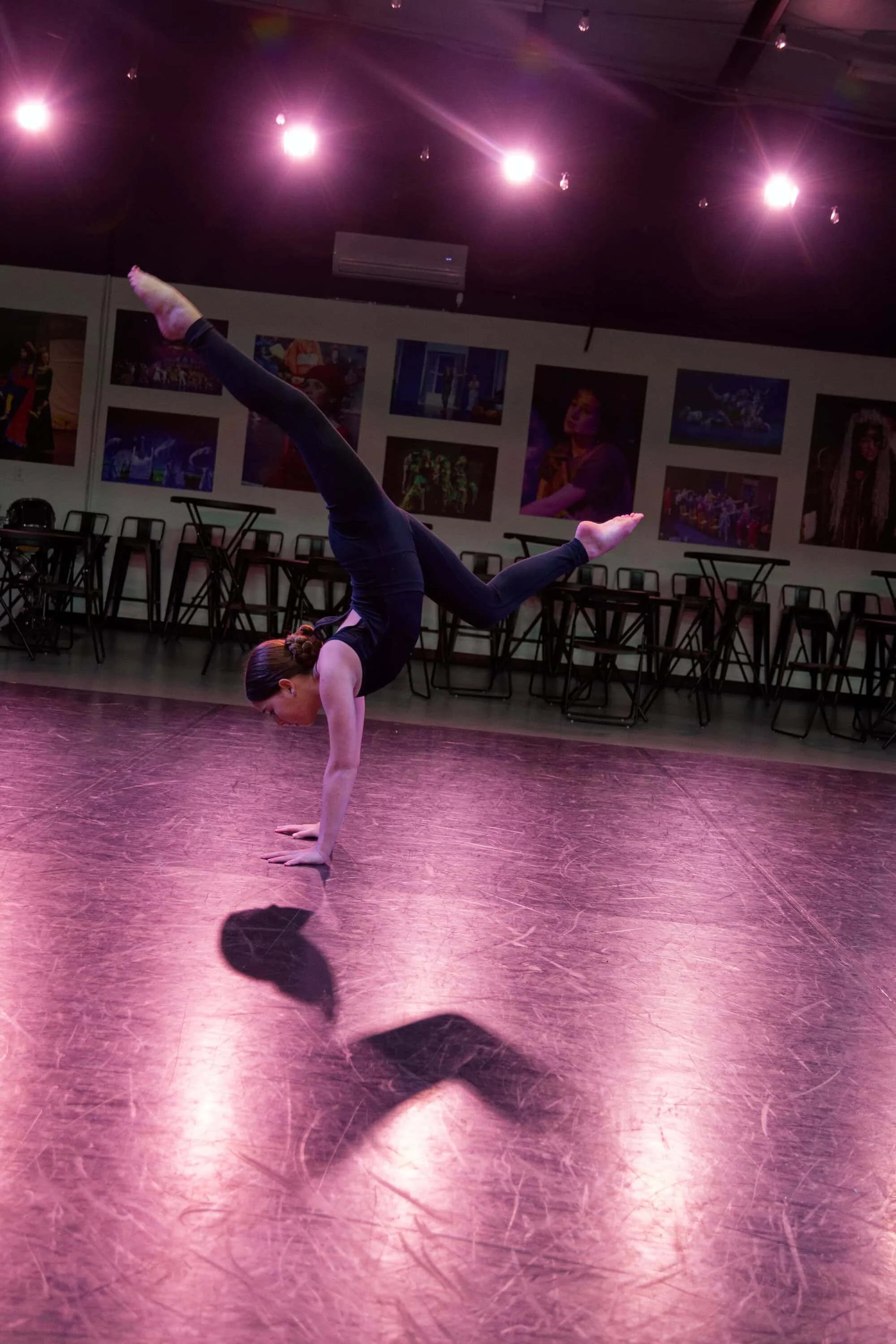 A woman performing a handstand in a dance studio with pink lighting and framed pictures on the wall.