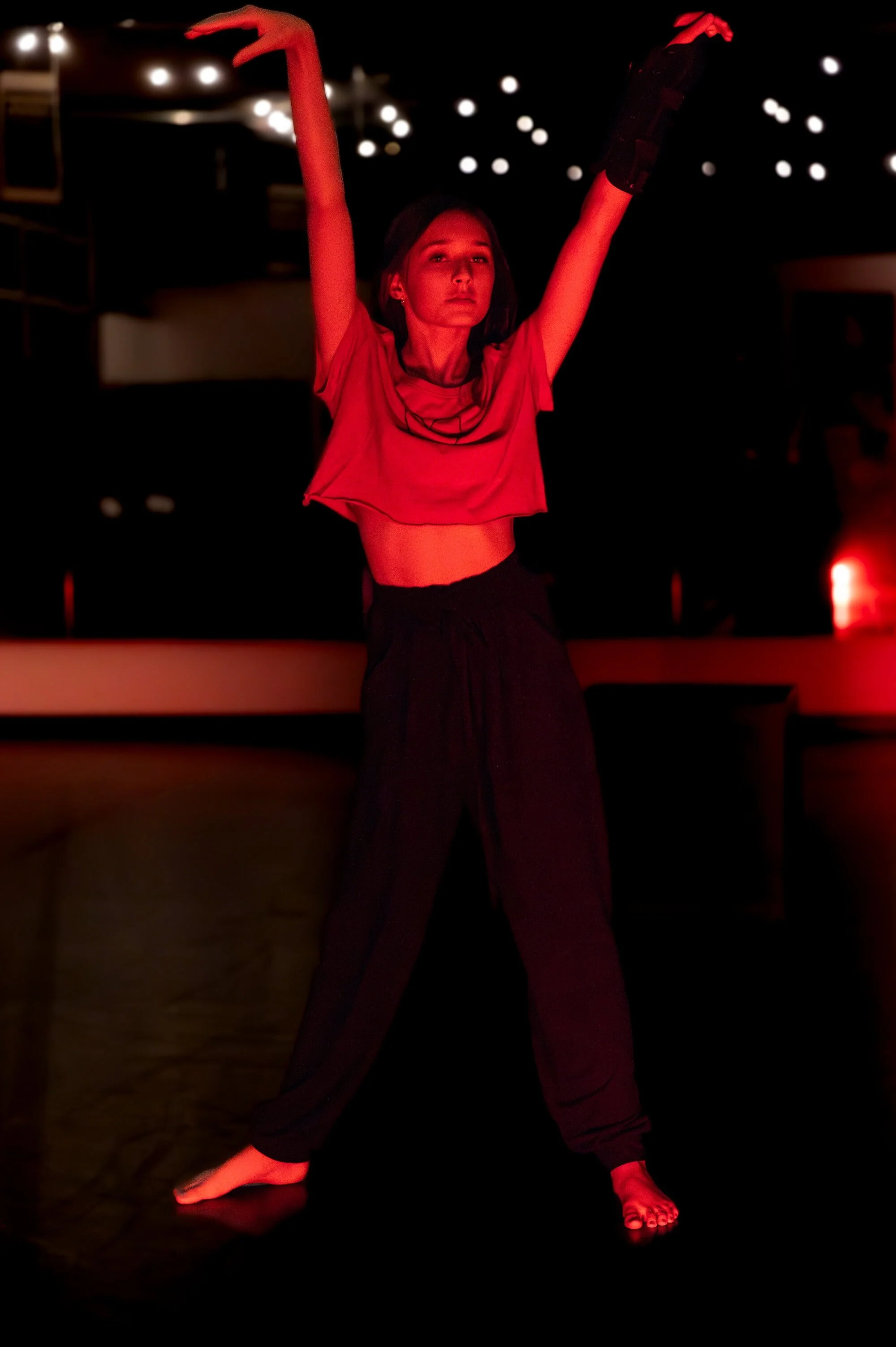 Young woman performing a dance move in a dark studio, illuminated by red stage lighting, with arms raised and dressed in a red crop top, black pants, and black gloves.