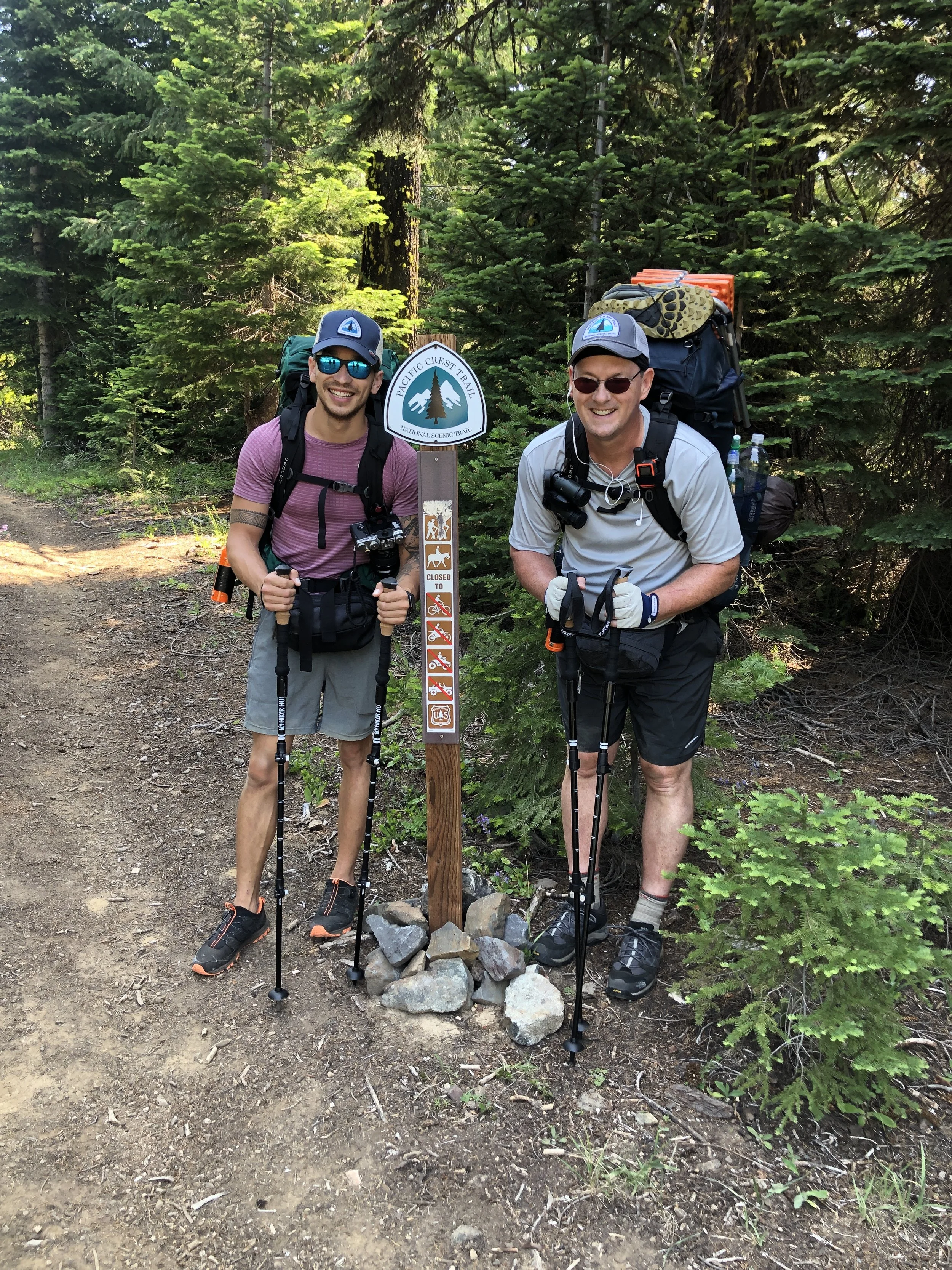 Two hikers standing on a forest trail, smiling and holding hiking poles, next to a trail sign that indicates the Pacific Crest Trail in a wooded area.