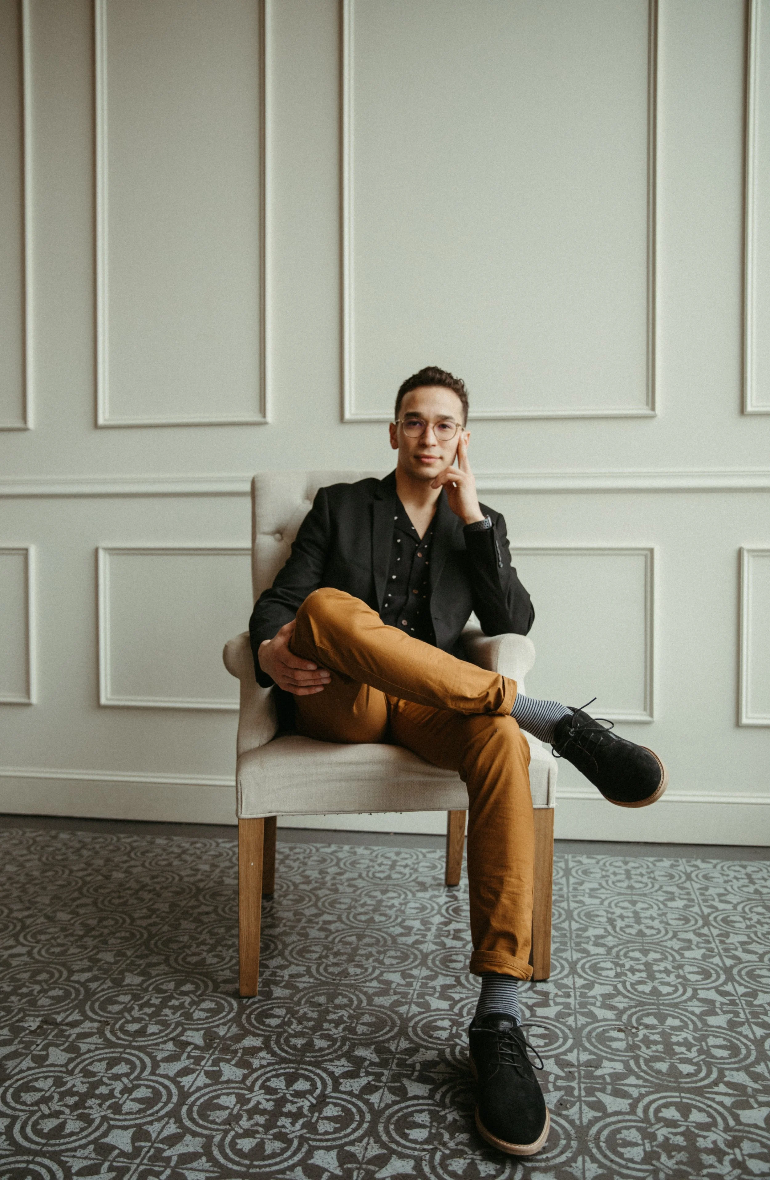 A young man sitting in an armchair in a well-lit room with decorative wall paneling and patterned tile flooring. He is wearing glasses, a black blazer, a black shirt, and tan pants, with one leg crossed over the other for Desert Dog Films Bend Oregon