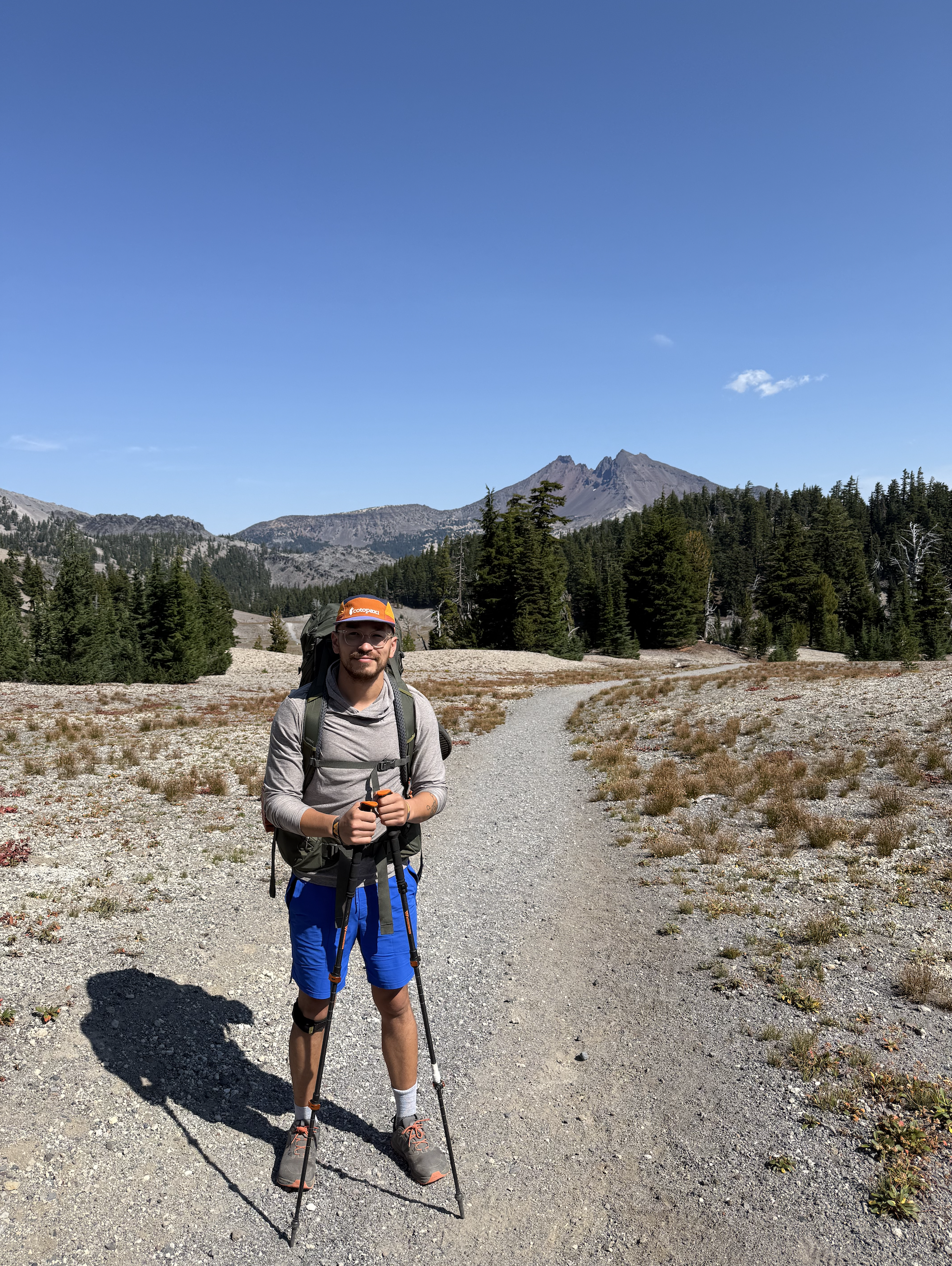 A man wearing a gray hoodie, blue shorts, and hiking boots stands on a trail in a mountainous area, holding trekking poles, with a backpack and sunglasses, under a clear blue sky, surrounded by trees and mountains for Desert Dog Films Bend Oregon