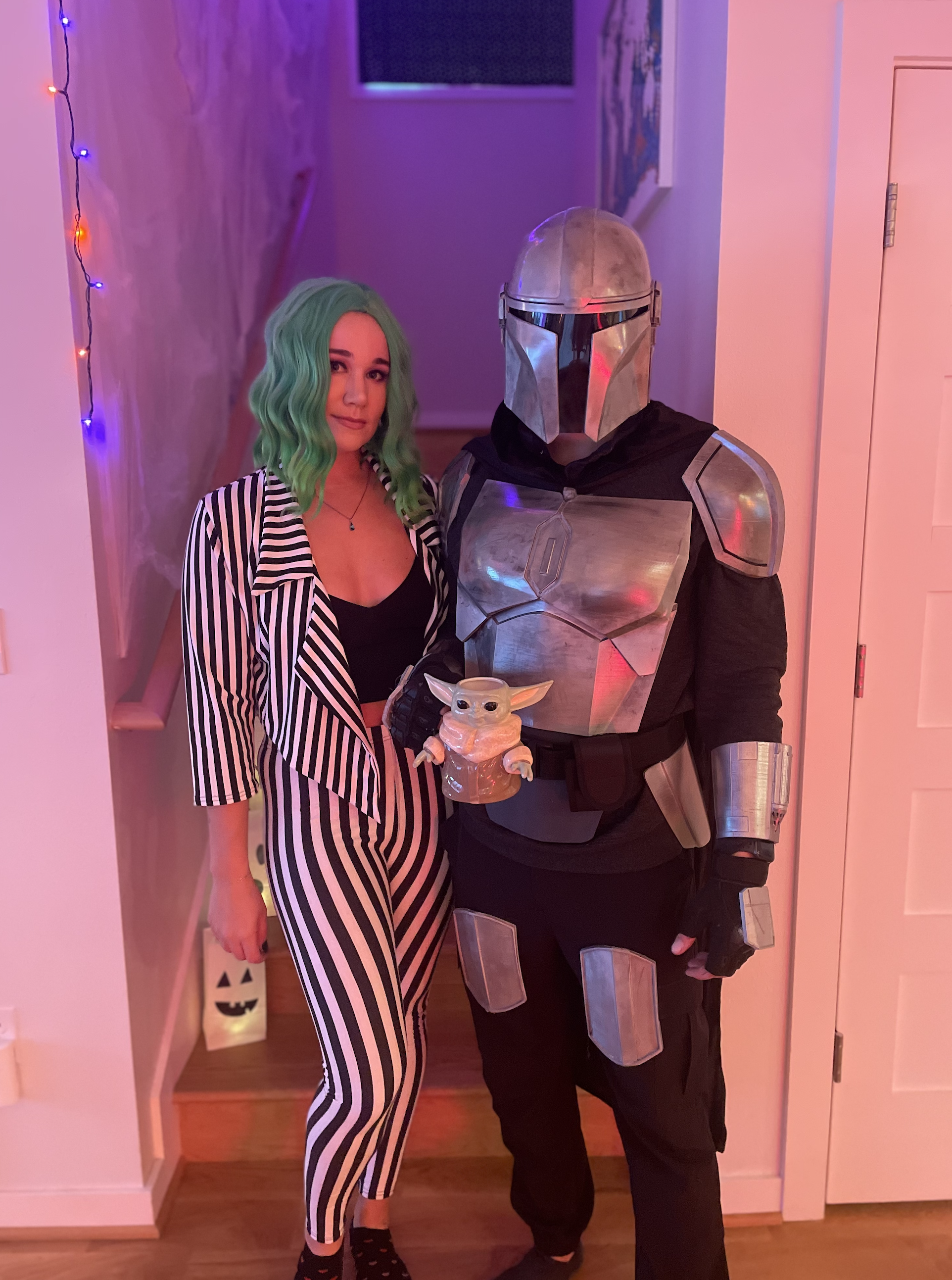 A woman with green wavy hair dressed in black and white striped pajamas and a man in Mandalorian costume with a Baby Yoda figure, celebrating Halloween in a decorated indoor setting with purple and pink lighting for Desert Dog Films Bend Oregon video