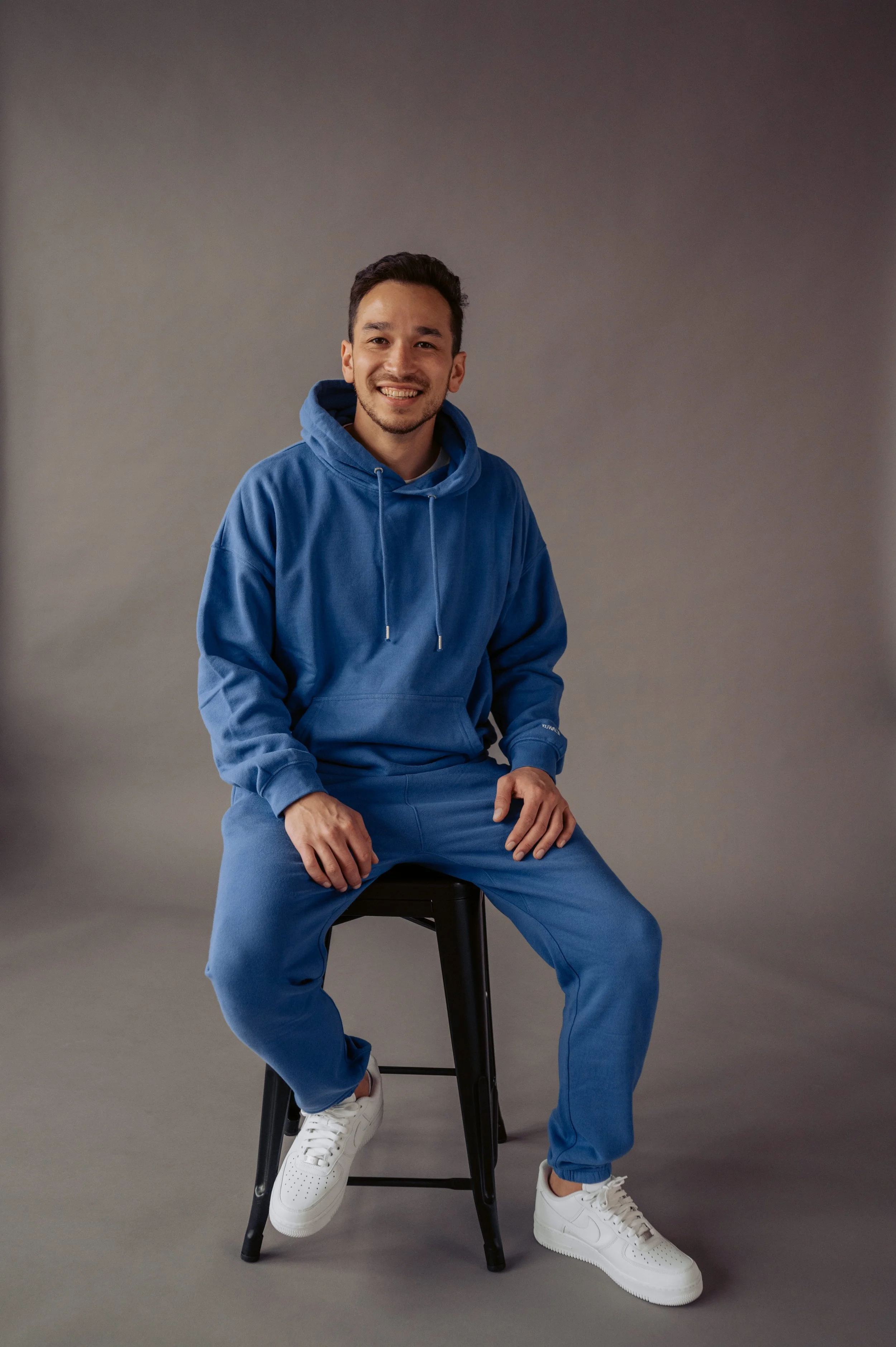 A man in a blue hoodie and sweatpants sitting on a black stool, smiling, against a plain background, wearing white sneakers for Desert Dog Films Bend Oregon video production company