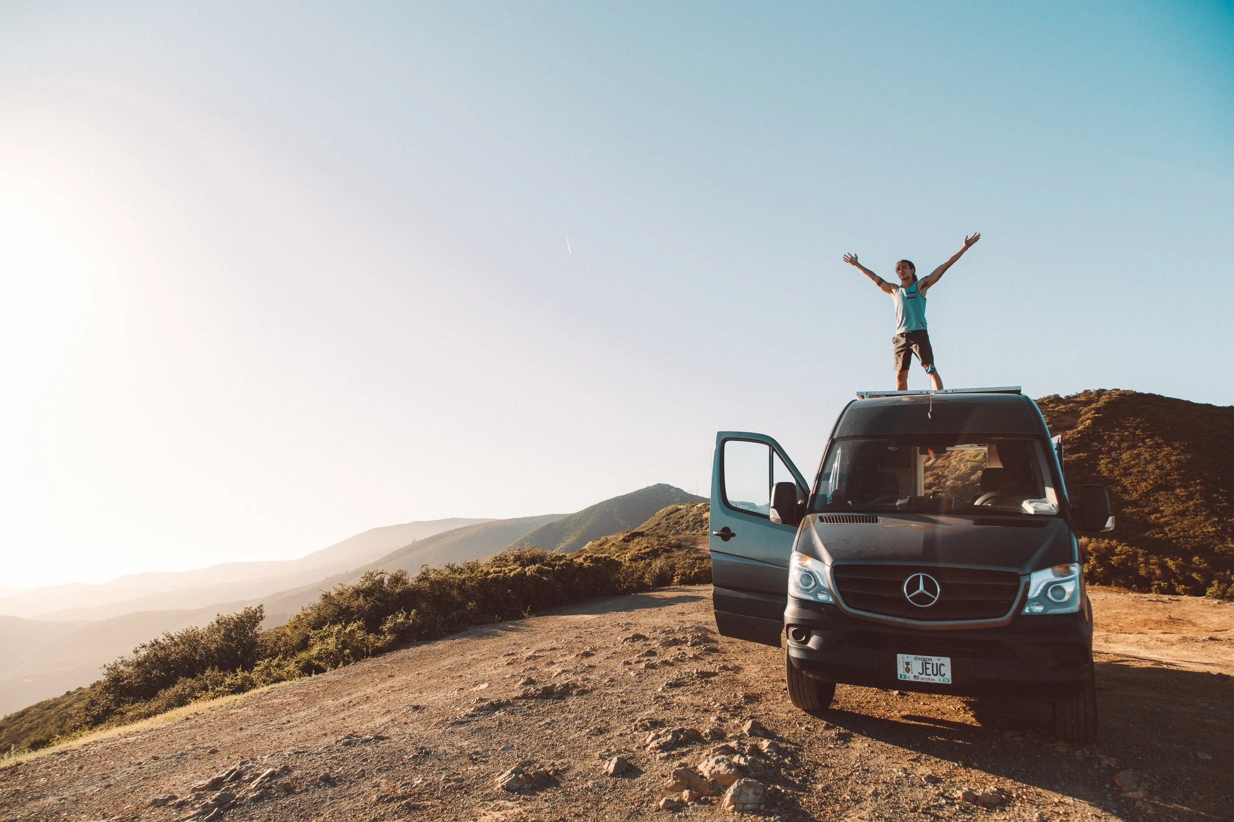 A person standing on top of a black Mercedes-Benz van with open passenger door, arms outstretched, on a dirt mountain trail at sunset with mountains and clear sky in background. for Desert Dog Films Bend Oregon video production company