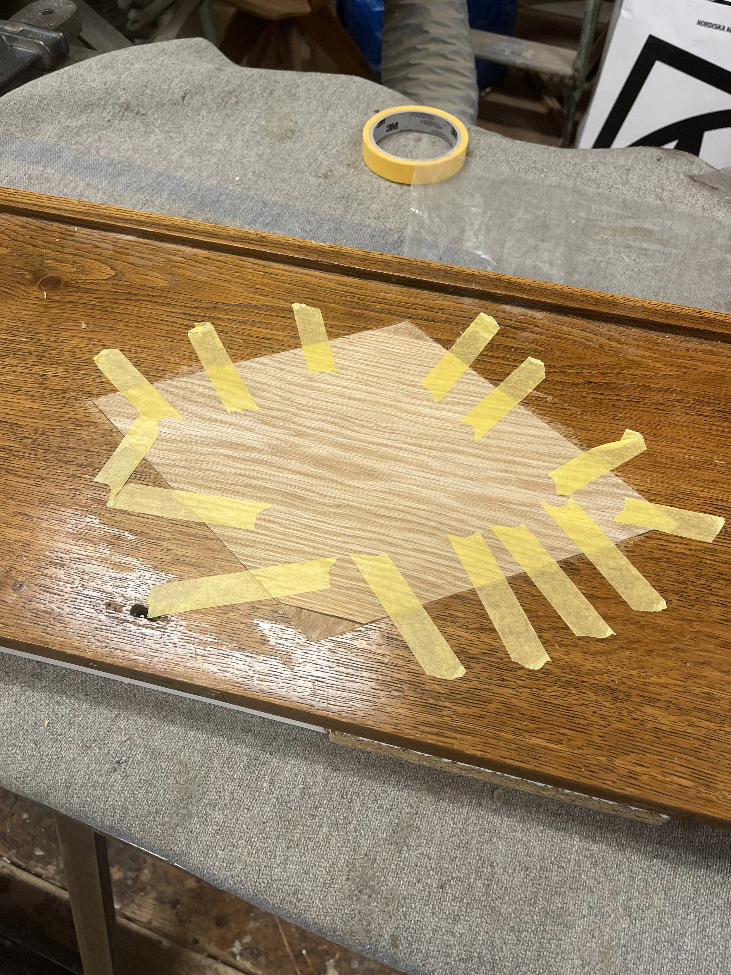 A woodworking workspace with a piece of wood taped with yellow painter's tape, possibly for marking or protection, on a wooden table, with torn masking tape and sawdust visible.