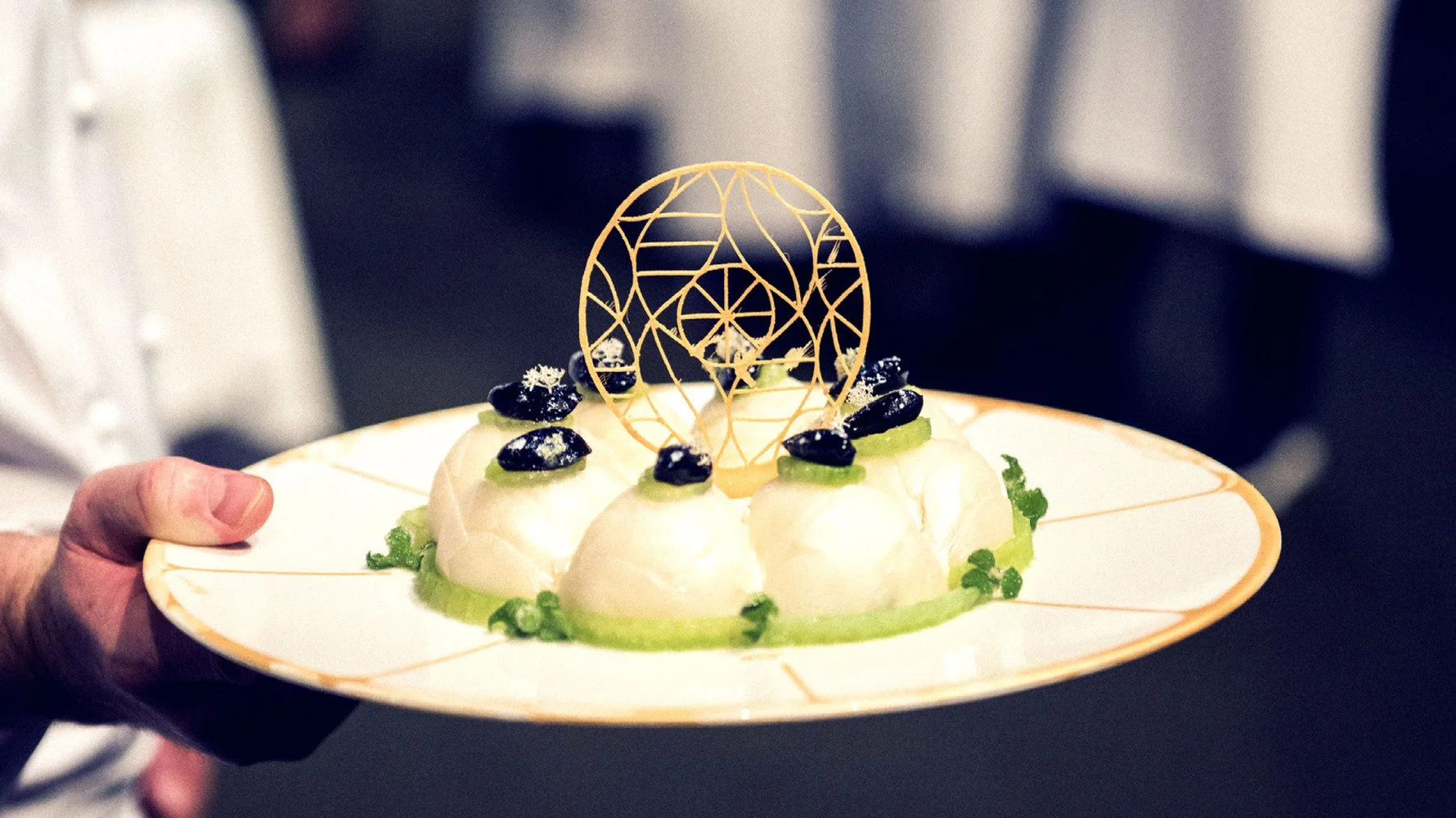 Chef holding a white plate with a dessert decorated with a gold geometric edible ornament, blackberries, and green garnishes.