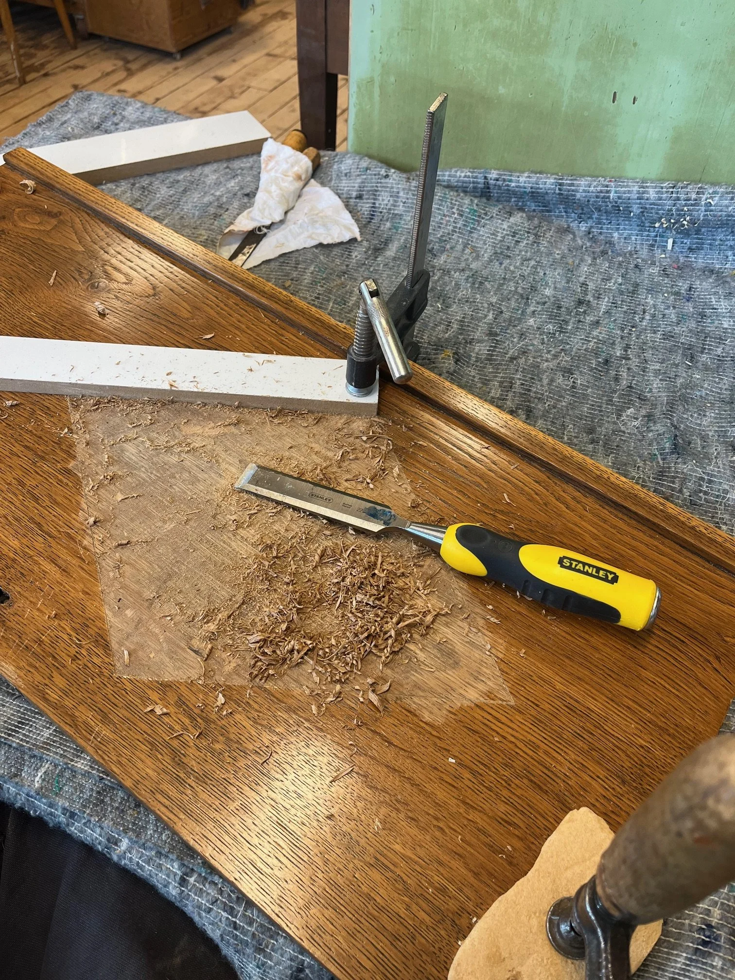 A woodworking workspace with a wooden table, a clamp securing a piece of wood, a utility knife, and wood shavings scattered on the surface.