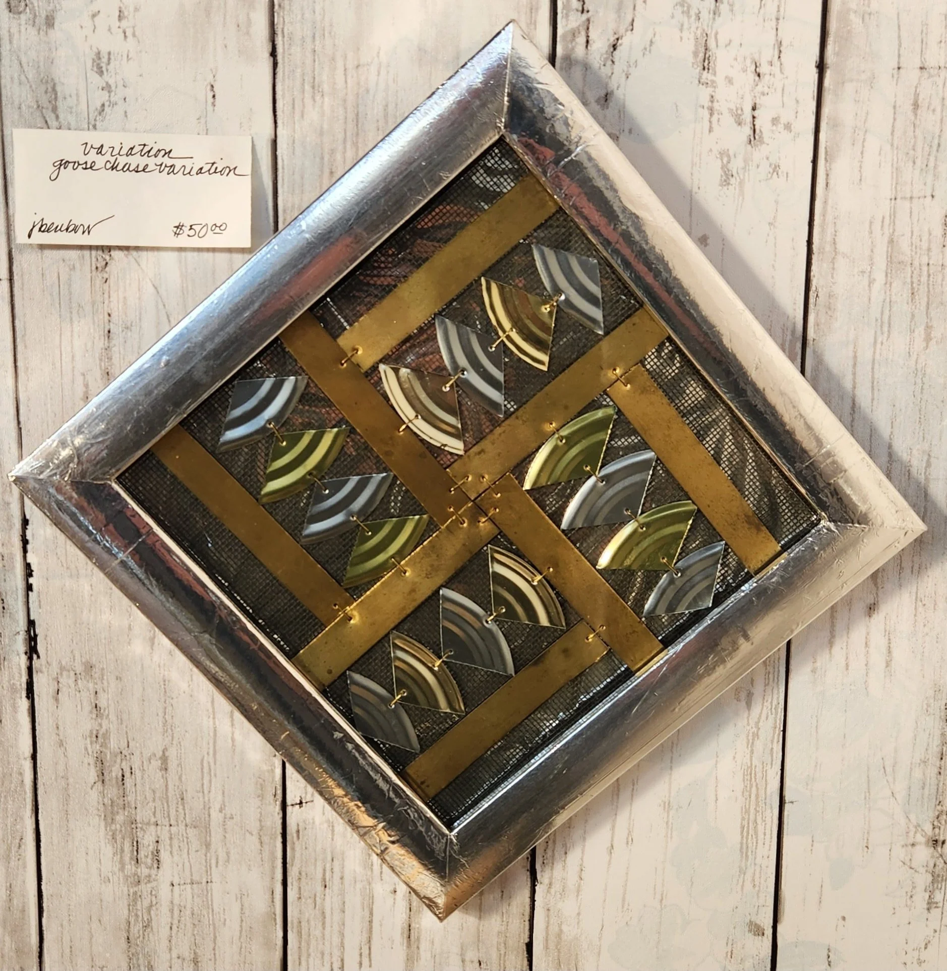 A framed art piece containing a pattern of black, white, and gold fan-shaped objects mounted on a wooden wall, with a handwritten price tag of $50.00.