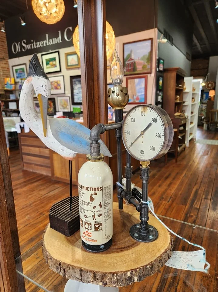 Decorative sculpture of a pelican with a fire extinguisher base, a pressure gauge, and a small lantern, mounted on a wooden slab in an art gallery.