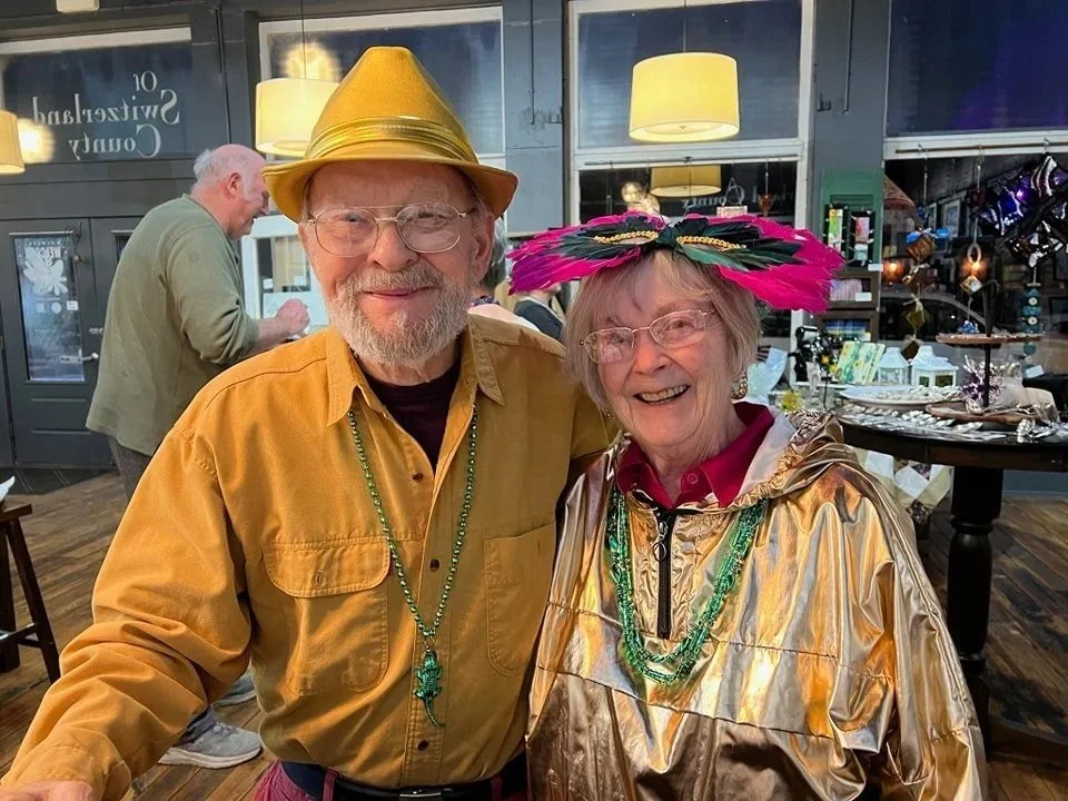 An elderly man and woman dressed festively at an indoor event, wearing hats and colorful necklaces, smiling at the camera.