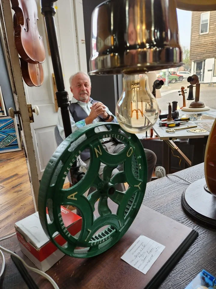 A man sitting indoors near a window, with vintage items including a green mechanical object, a visible lightbulb, and various small items on the table behind him, as seen through a foreground of hanging objects.