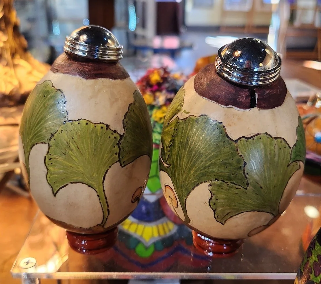 Two decorative egg-shaped objects with painted green leaves and a ceramic base, topped with shiny silver lids.